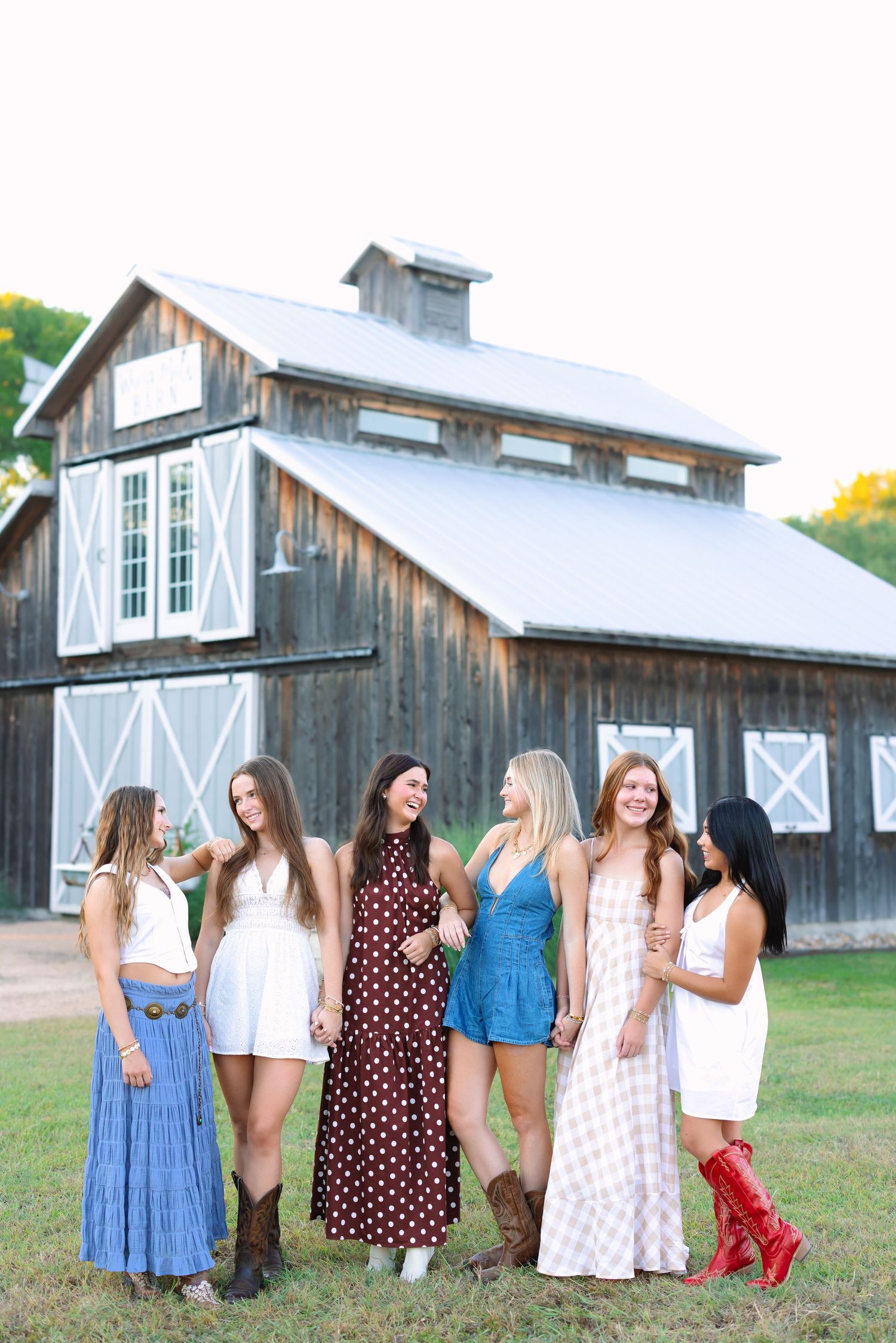 A group in sundresses and cowboy boots poses together in front of a rustic barn with white trim.