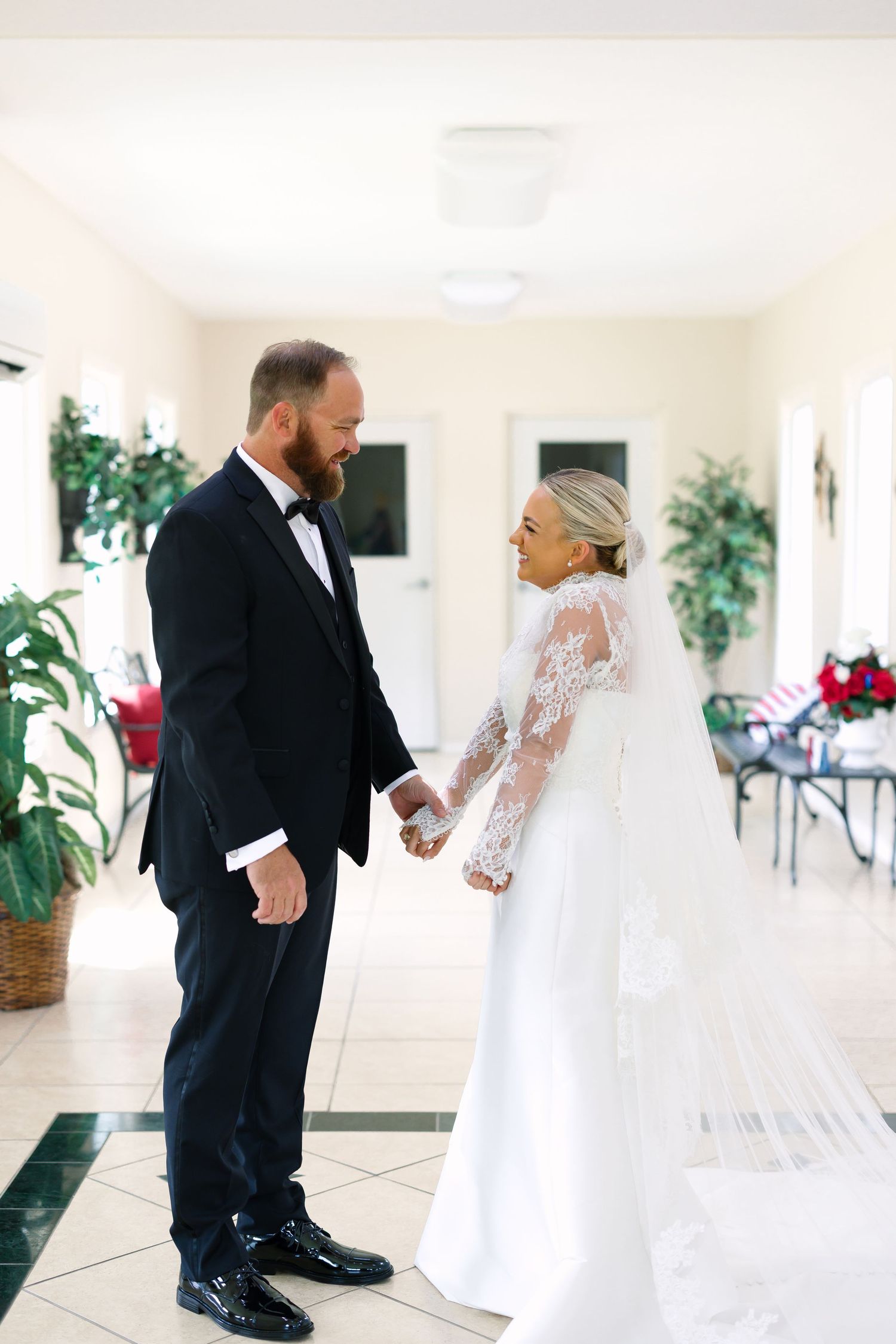 Bride in lace wedding gown with veil holds hands with bearded groom in black tuxedo indoors.