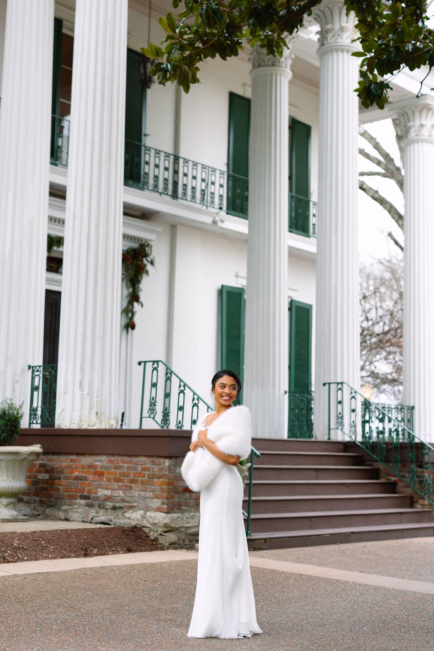 A woman in a white gown and fur wrap poses before a grand Southern mansion with white columns and green shutters.