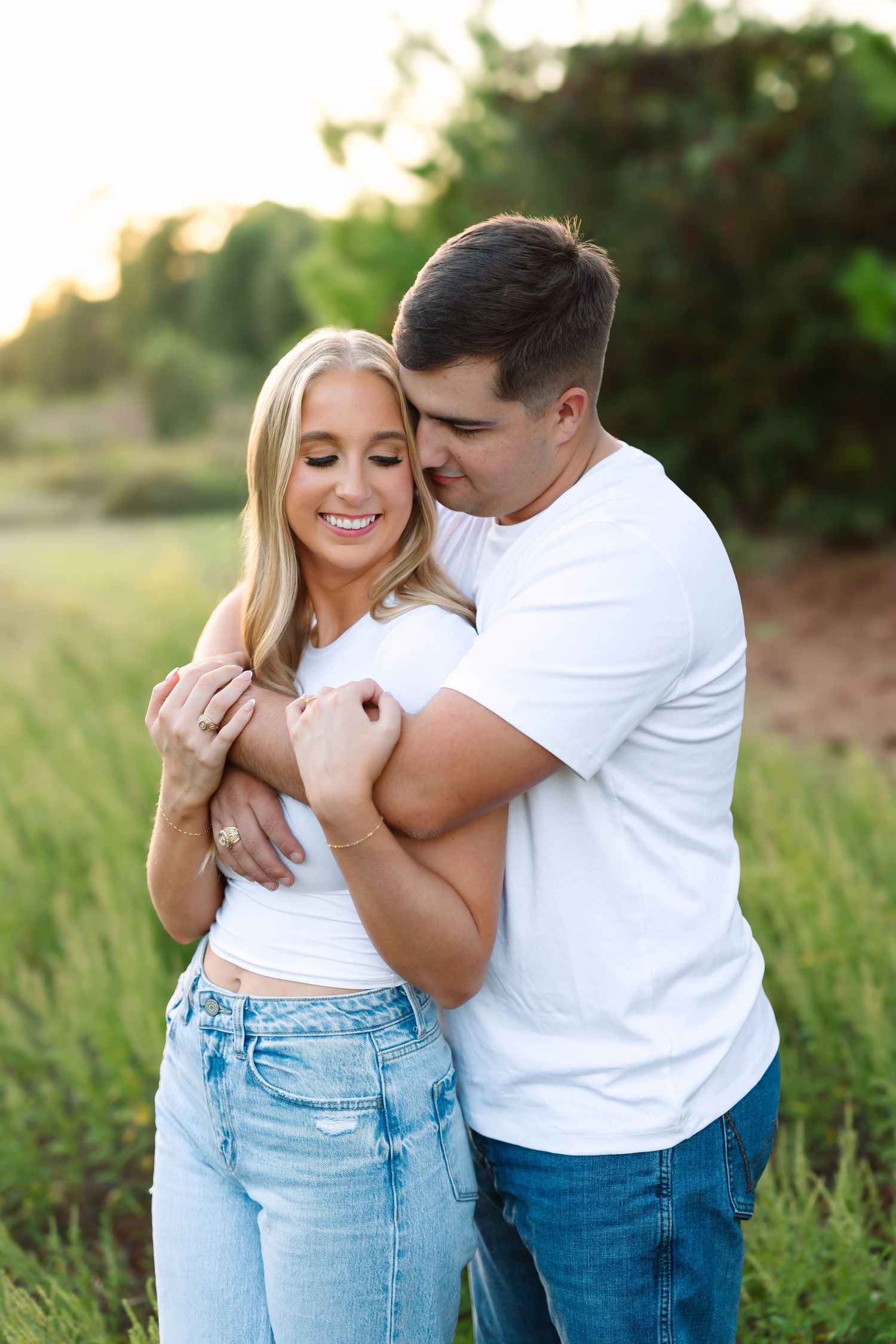 Couple embracing in golden hour light, man in white tee hugging woman in white crop top and jeans in green field.