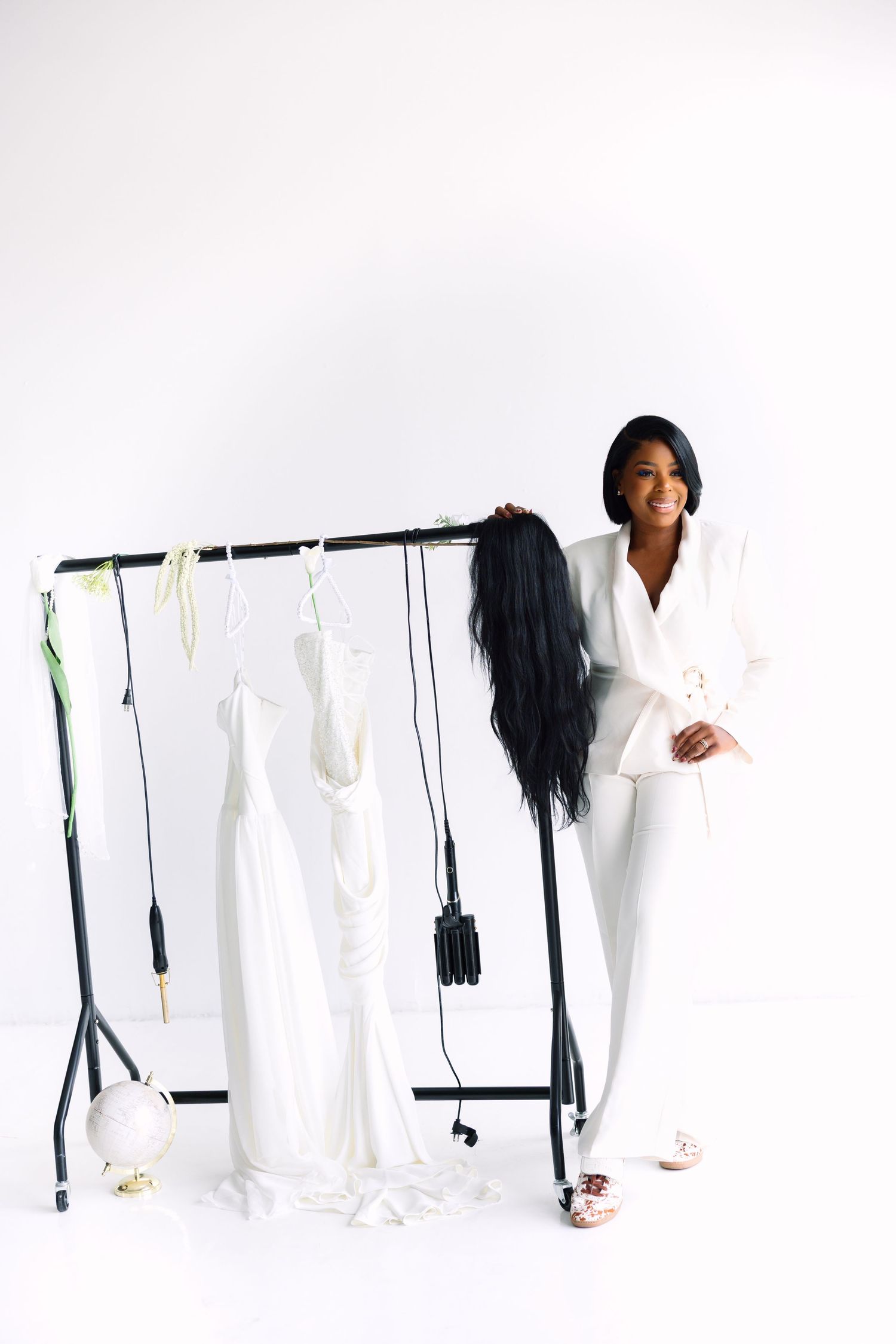 A smiling woman in a white suit stands beside a clothing rack with white dresses on a white background.