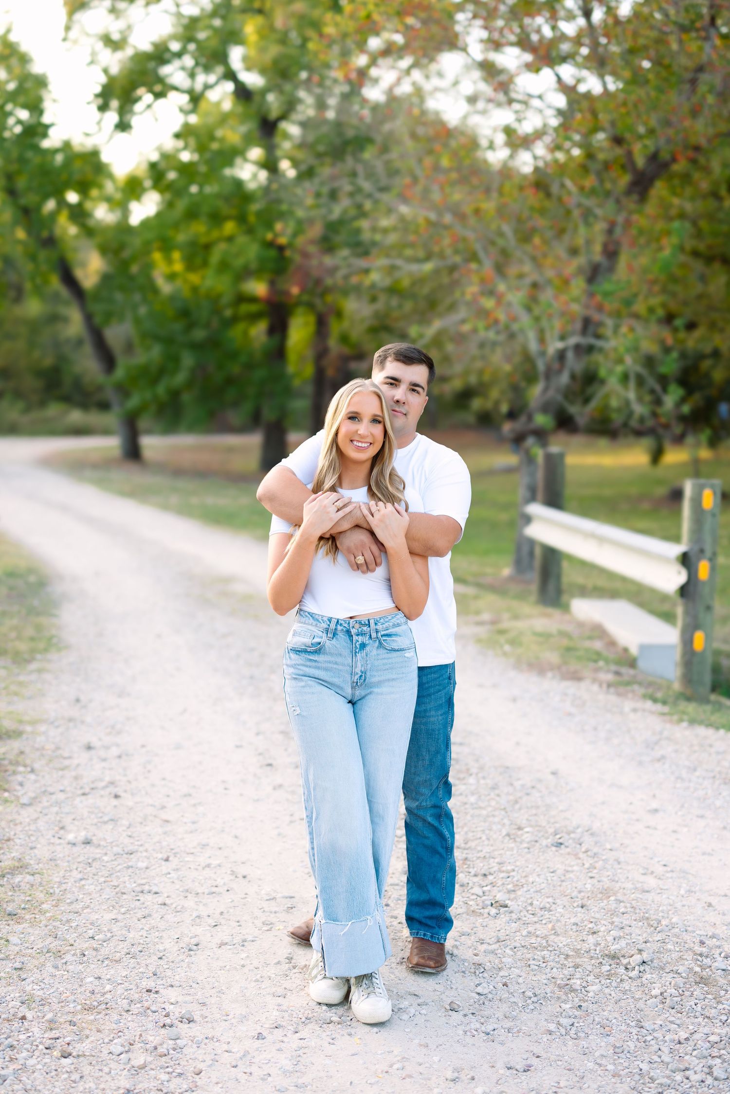 Couple in matching white tees and jeans embrace on a scenic gravel path surrounded by lush green trees.