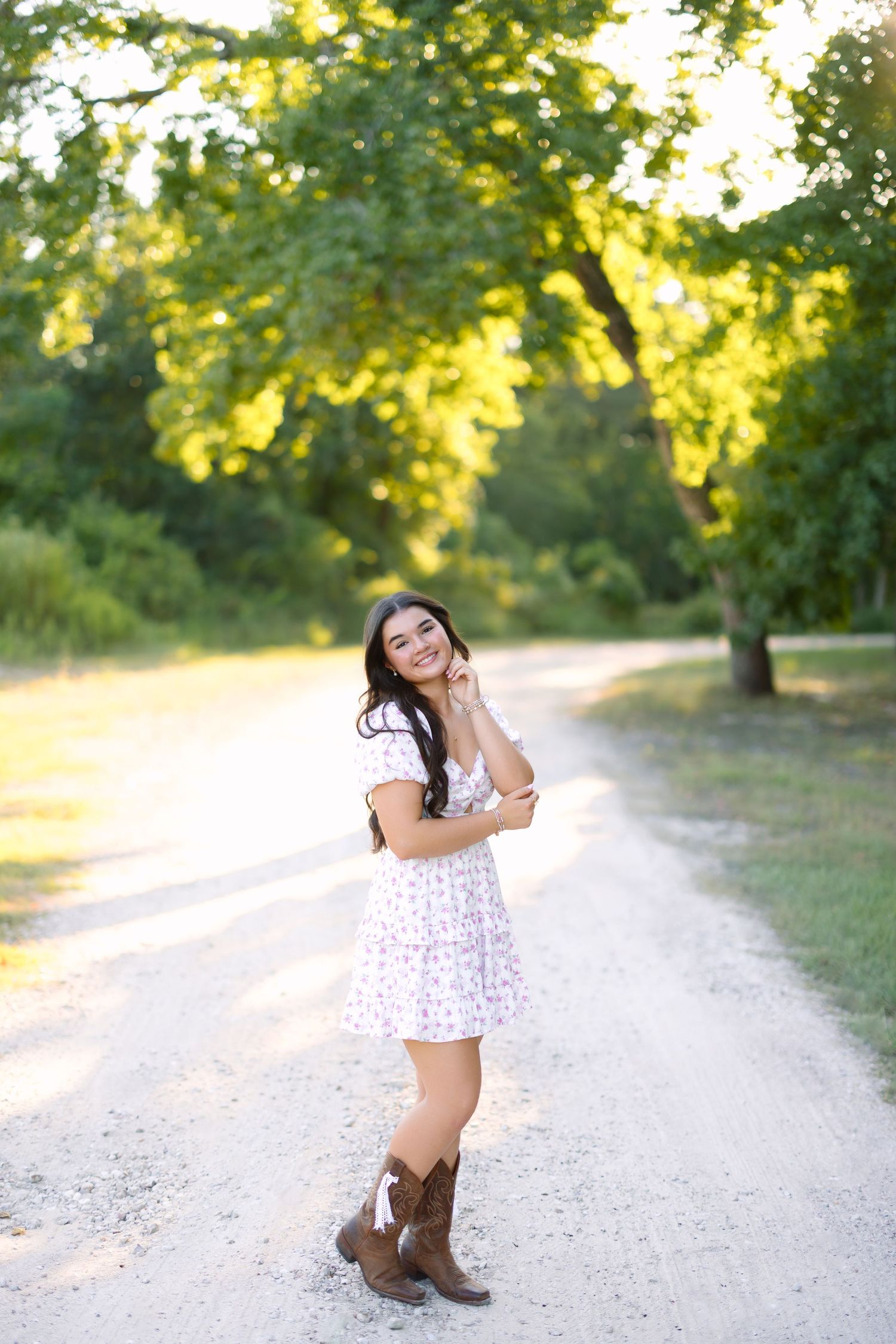 Young woman in floral dress and cowboy boots smiling on a sunlit dirt path surrounded by lush green trees.