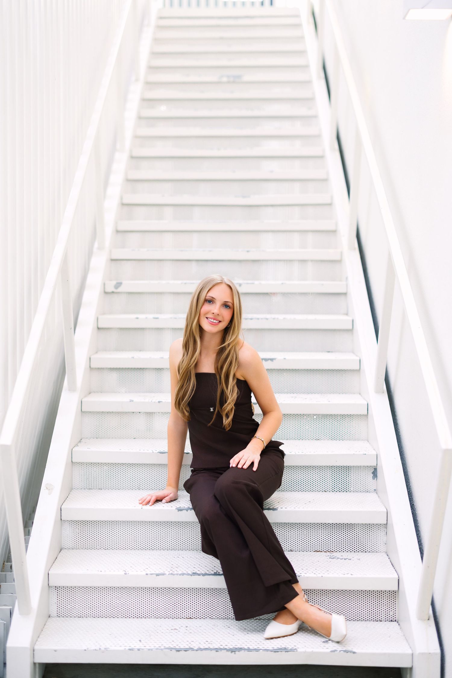 A smiling woman with long blonde hair sits on white outdoor stairs wearing a dark brown jumpsuit and white flats.