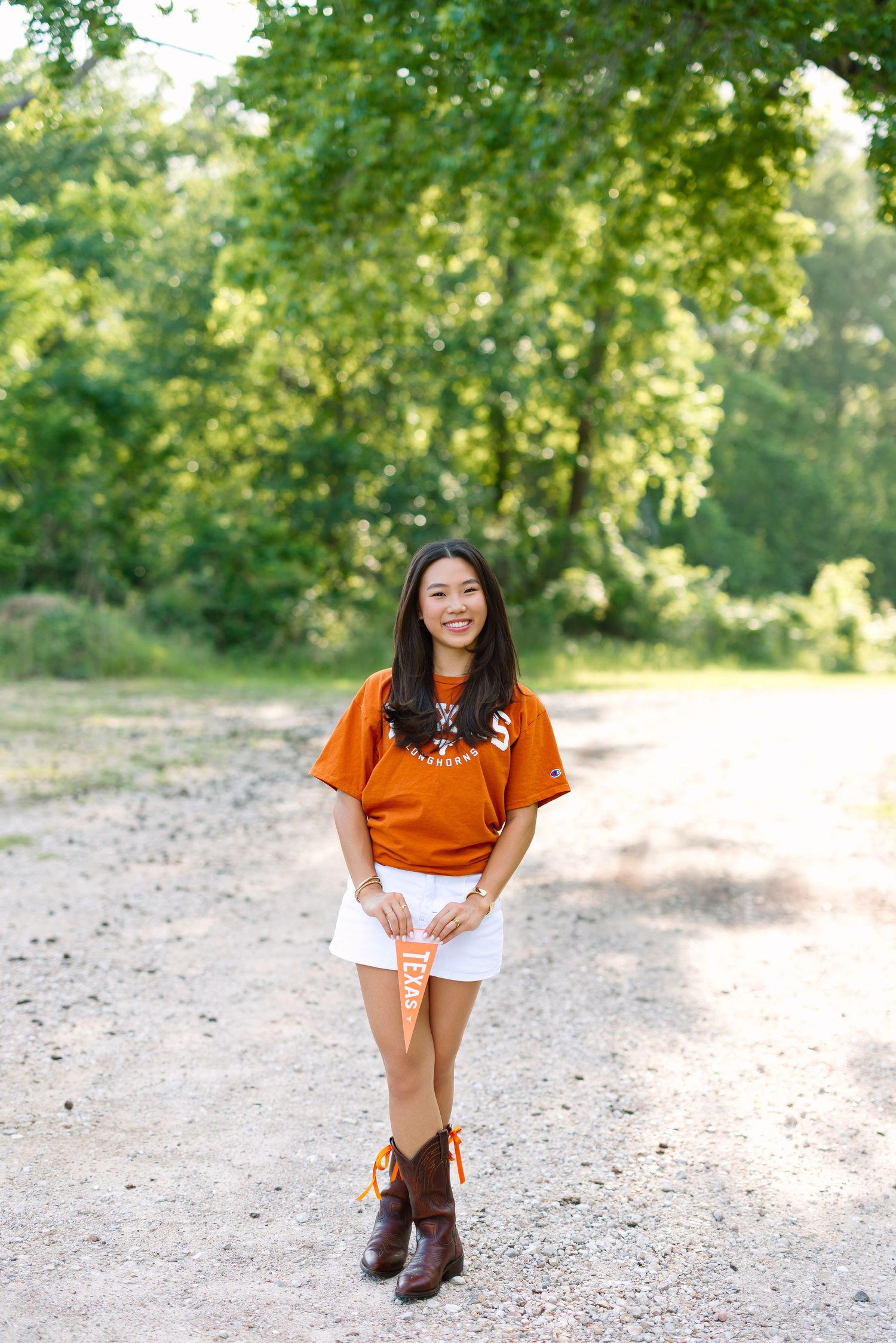 Young woman in orange Texas Longhorns shirt, white skirt, and cowboy boots holding a pennant on a gravel path.
