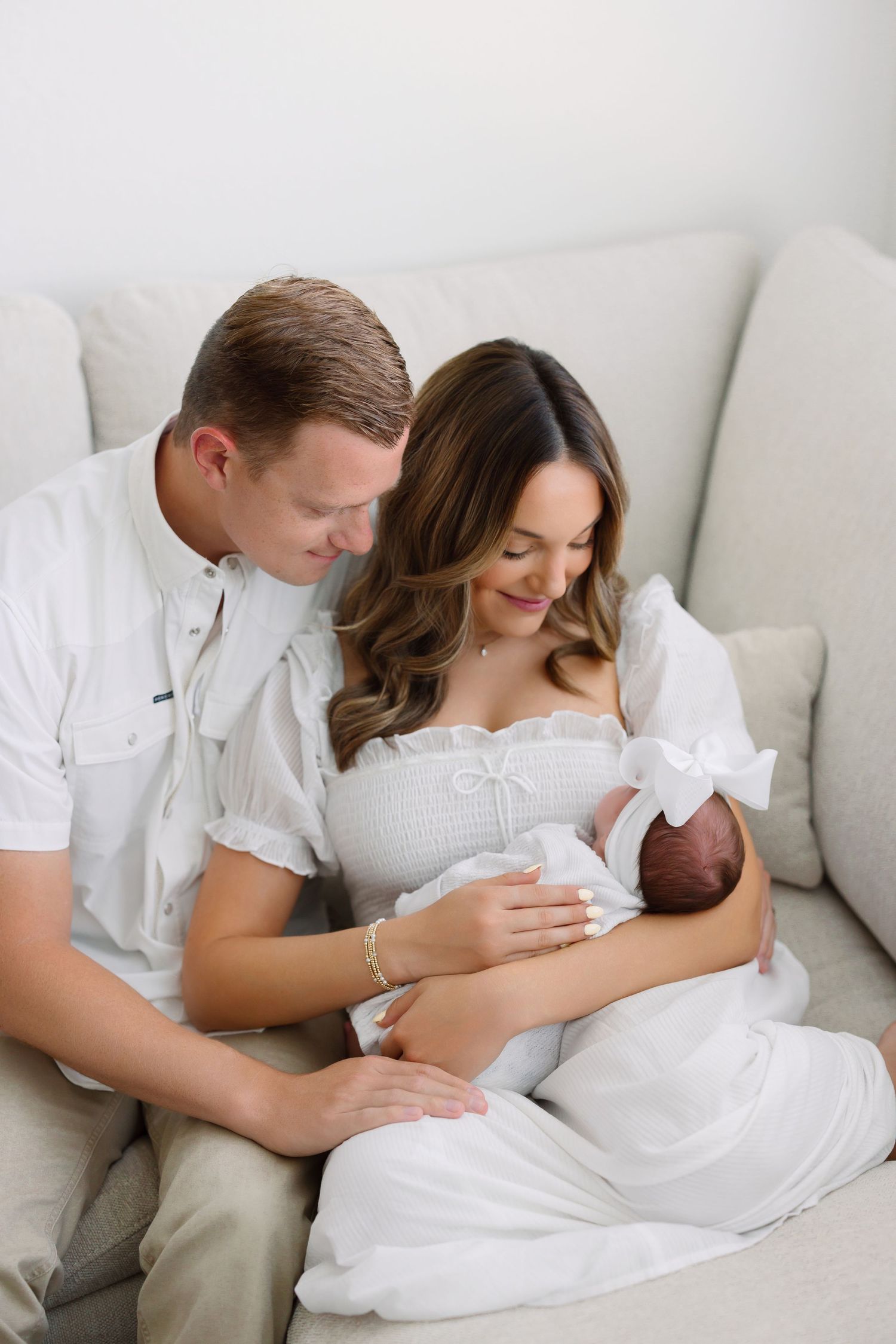 A couple in white outfits sits on a beige sofa, lovingly gazing at their newborn baby wrapped in white.