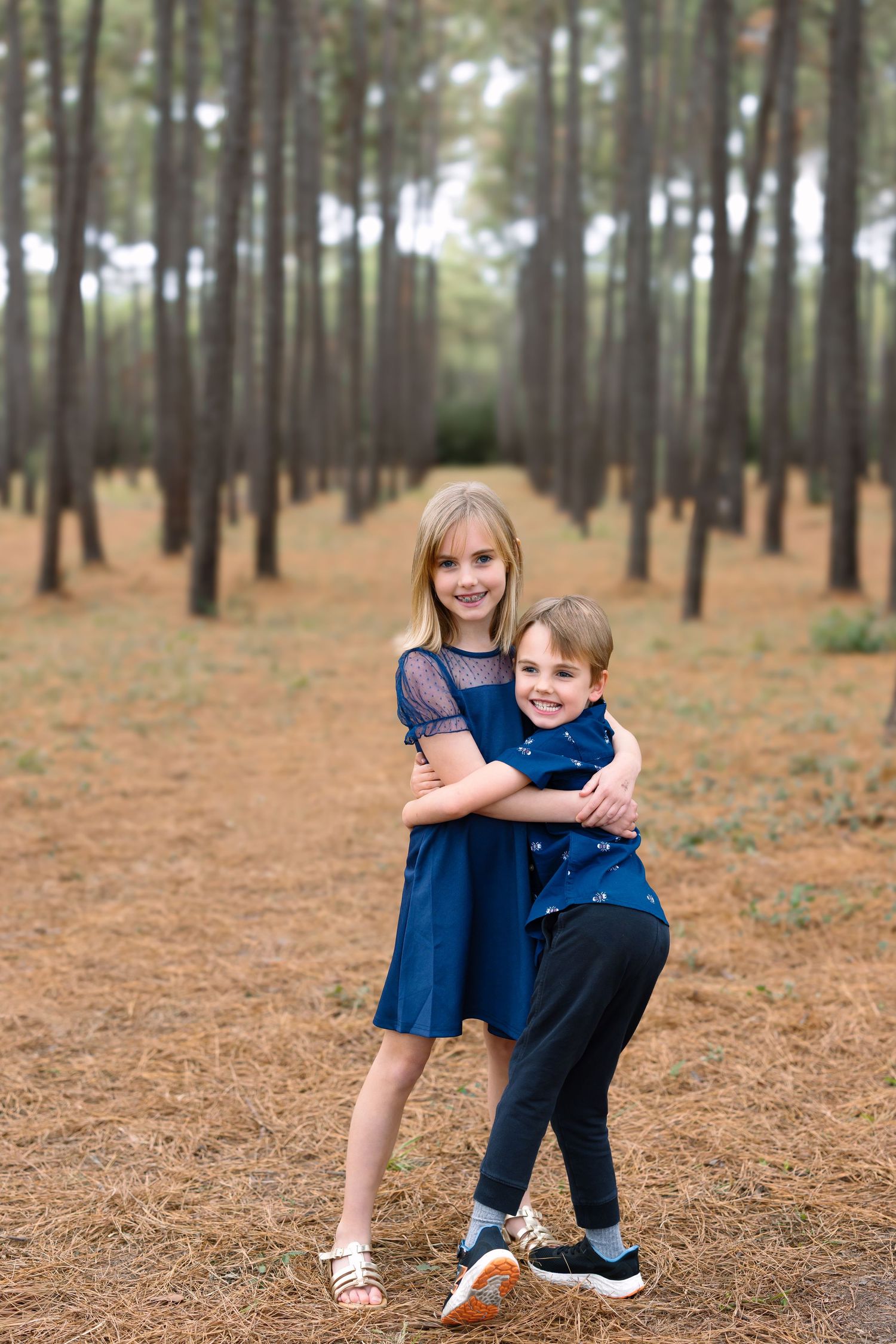 Two children in blue outfits hugging in a pine forest with rows of tall trees and golden fallen needles.