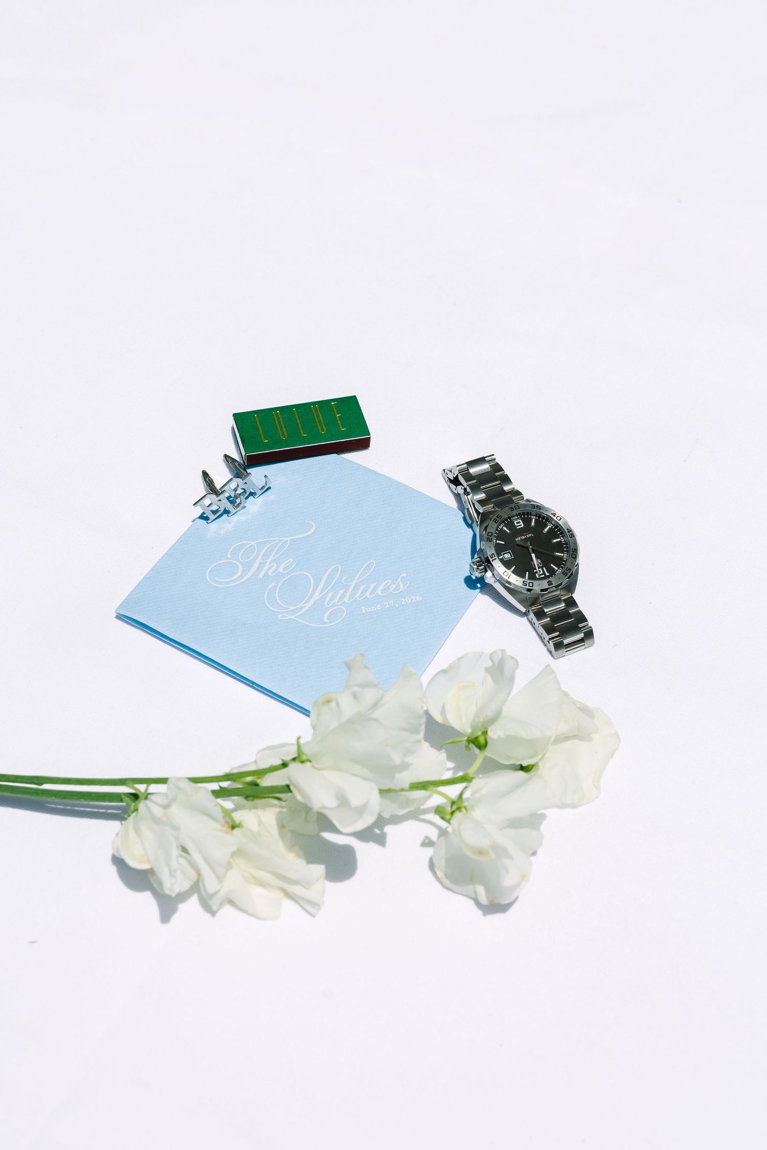 Groom accessories flat lay featuring silver watch, cufflinks, blue card, and white sweet pea flowers.