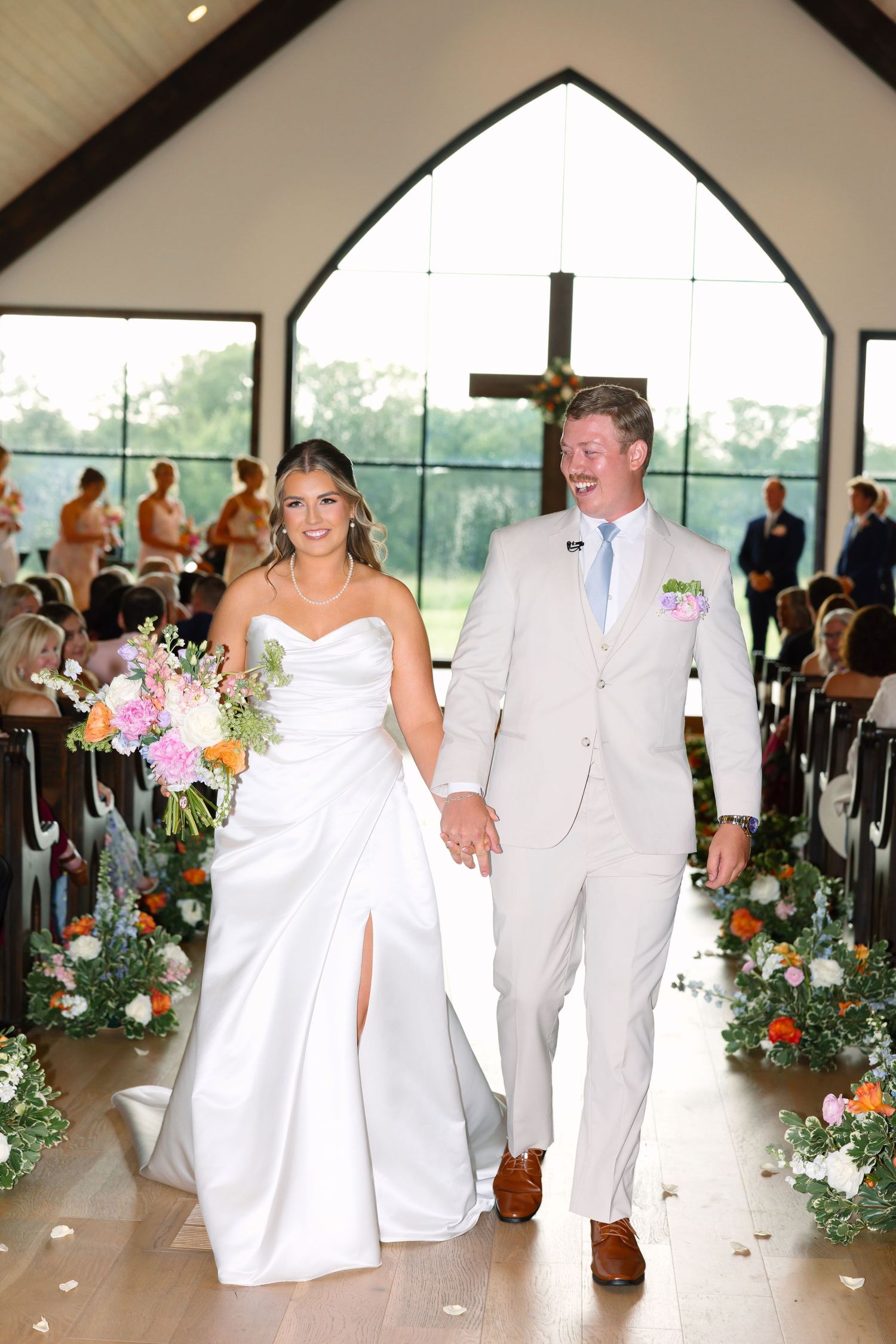 Smiling newlyweds walk down the aisle of a white chapel decorated with colorful floral arrangements.