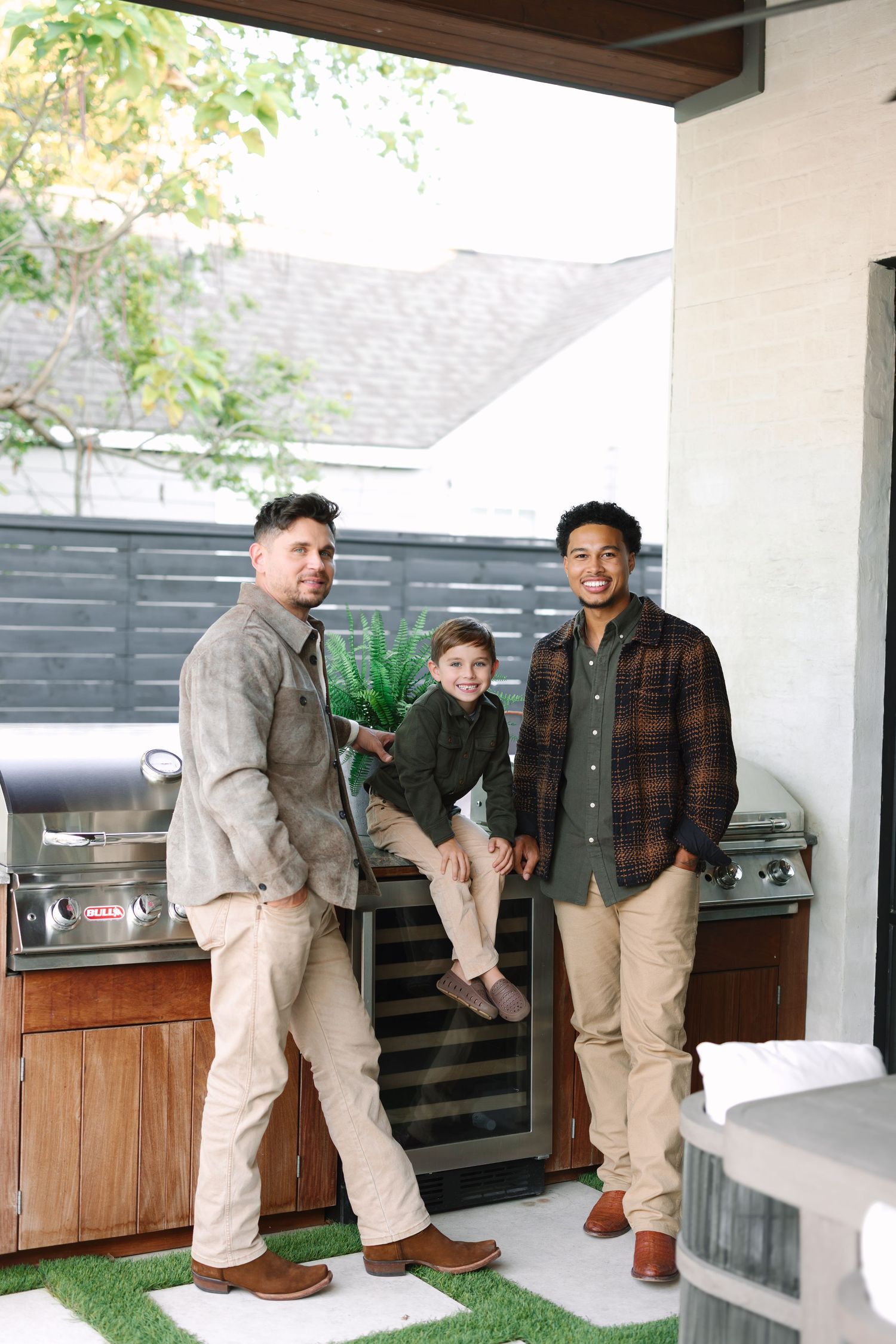 Two men and a young boy pose together in an outdoor kitchen area, all wearing casual earth-tone outfits.