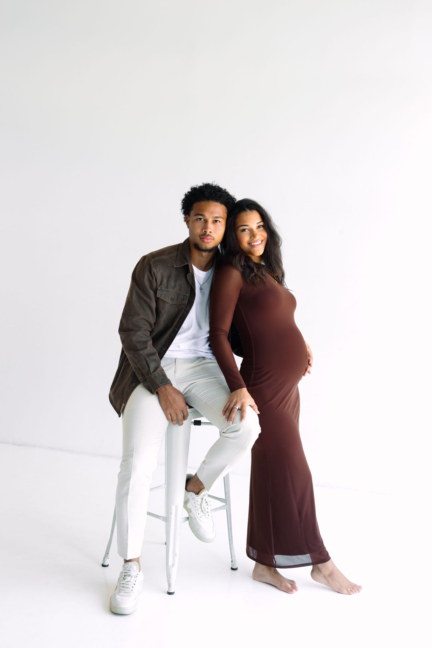 A man in a brown jacket sits on a stool beside a pregnant woman in a brown dress against a white background.