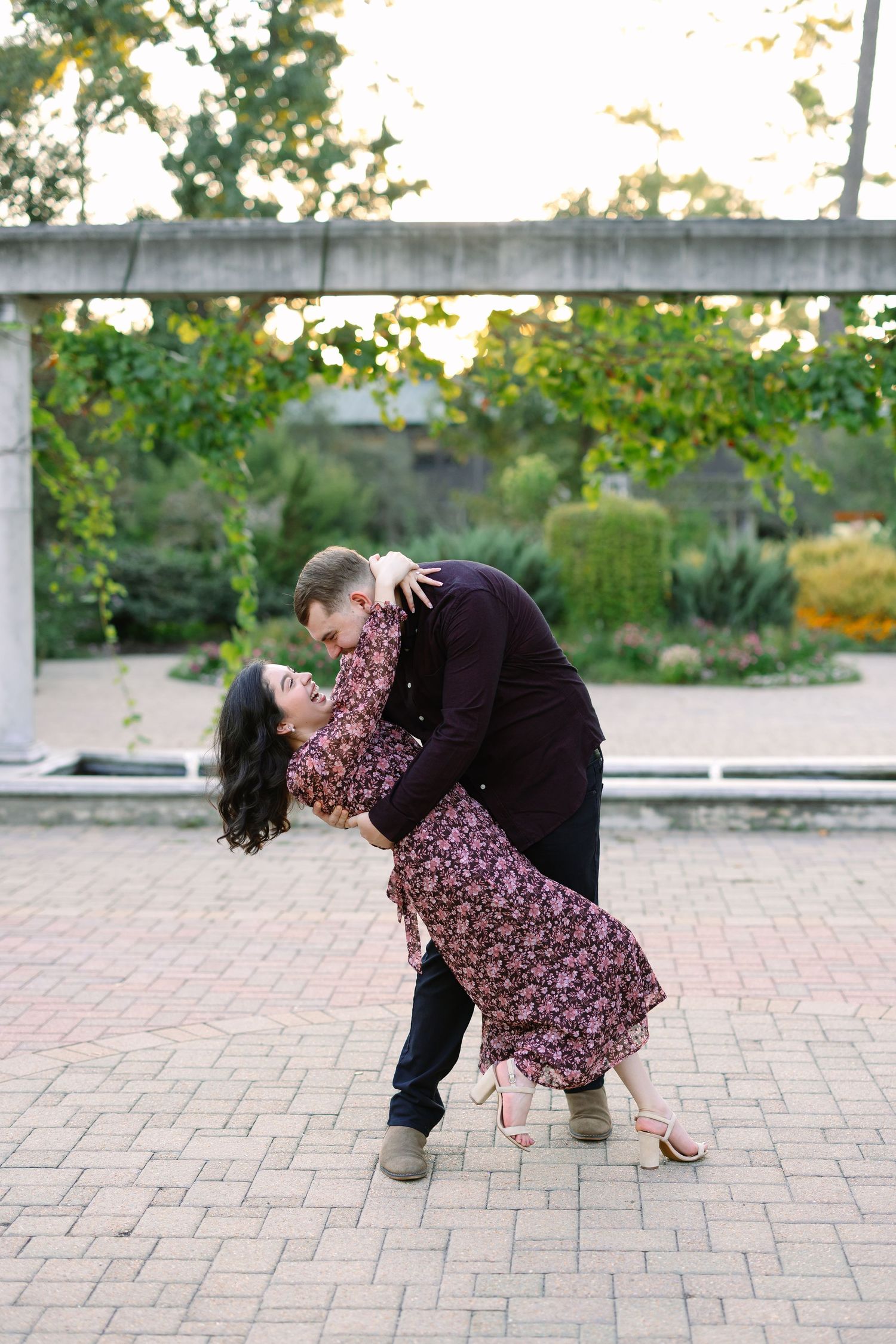 Couple sharing a romantic dip and near-kiss on a brick patio surrounded by greenery during a golden hour engagement session.