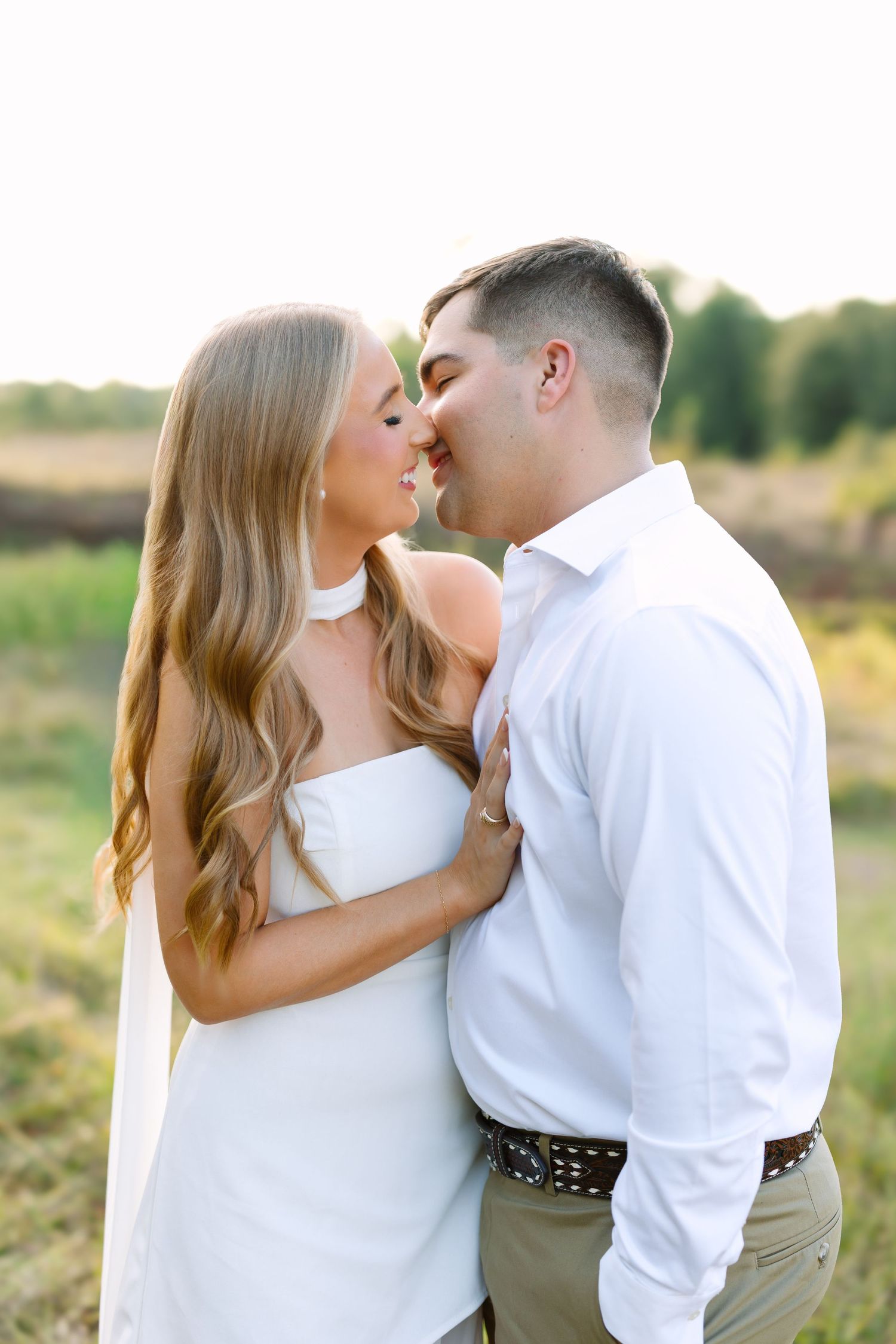 Couple sharing a romantic almost-kiss in a sunlit field, woman in white dress, man in white shirt.