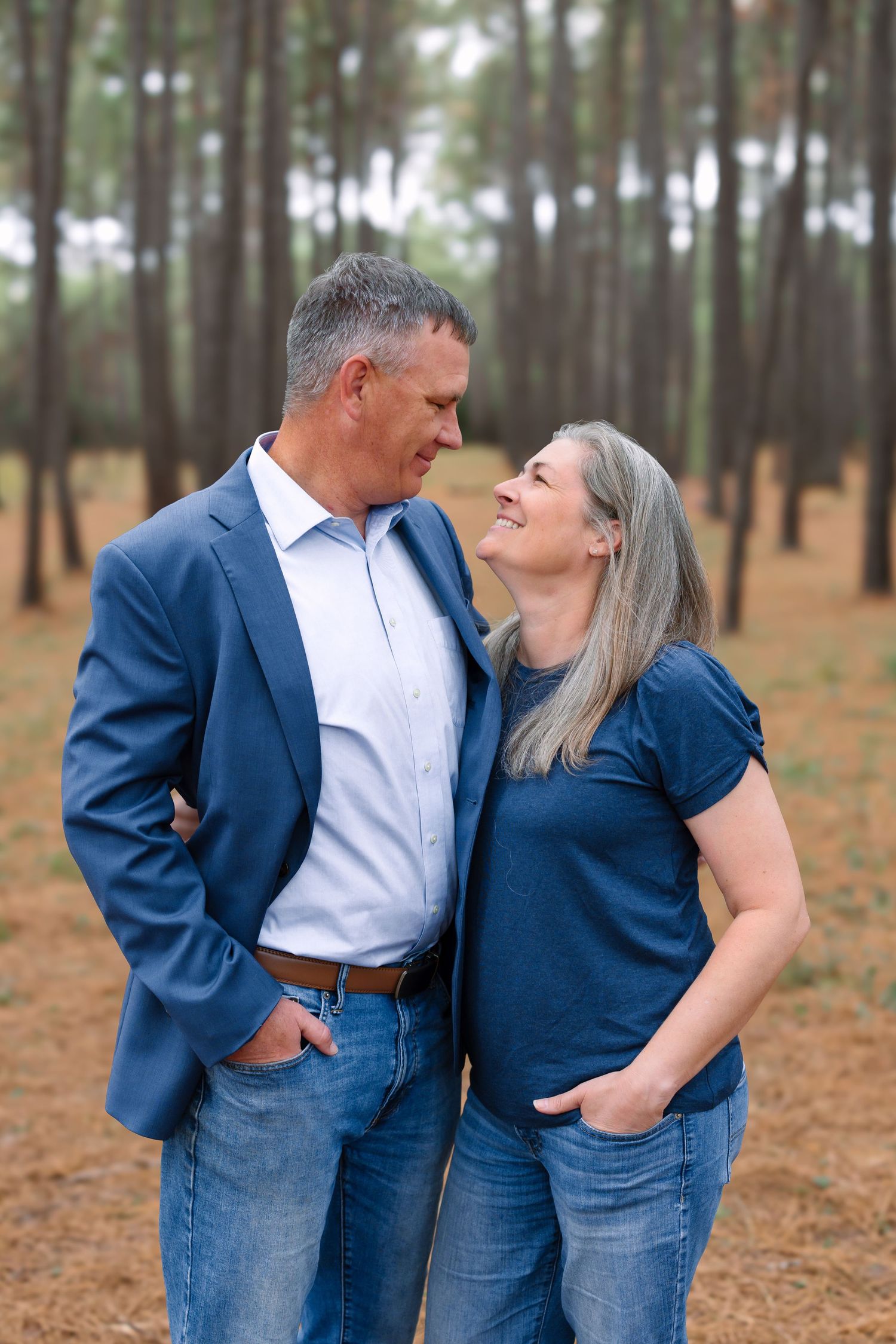 A mature couple in blue attire share a loving glance while standing in a pine tree forest.