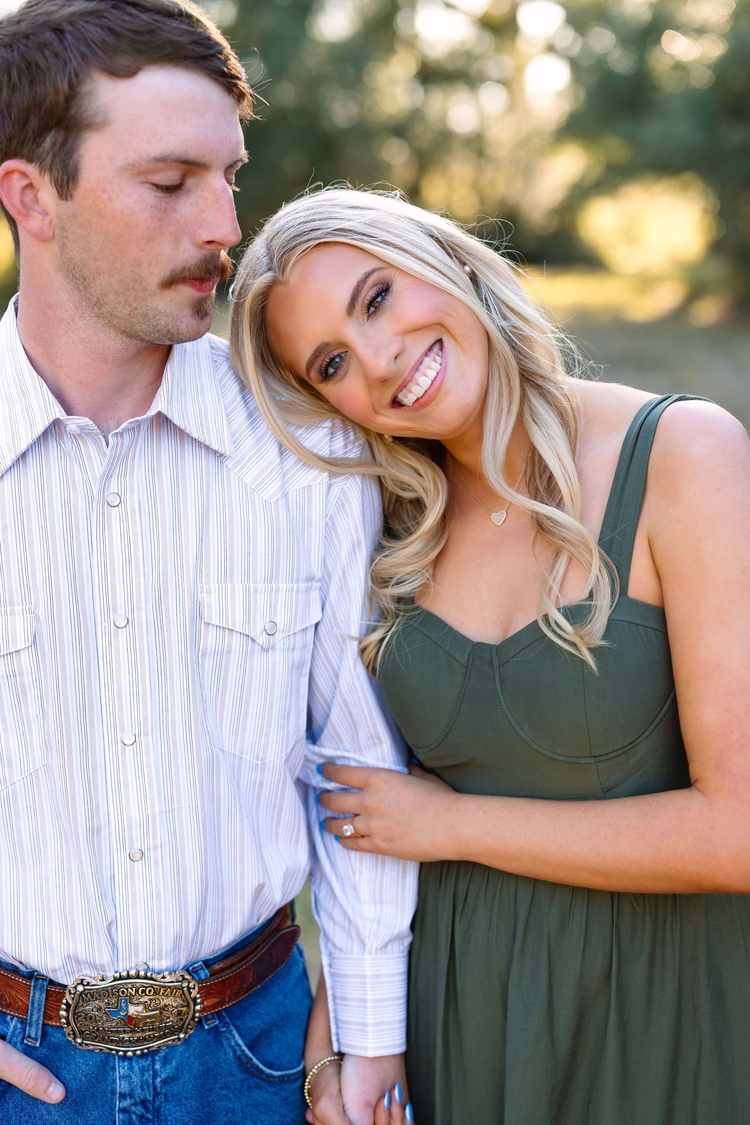 Couple in outdoor engagement photo, woman in green dress leans on man in striped shirt and jeans with western belt buckle.