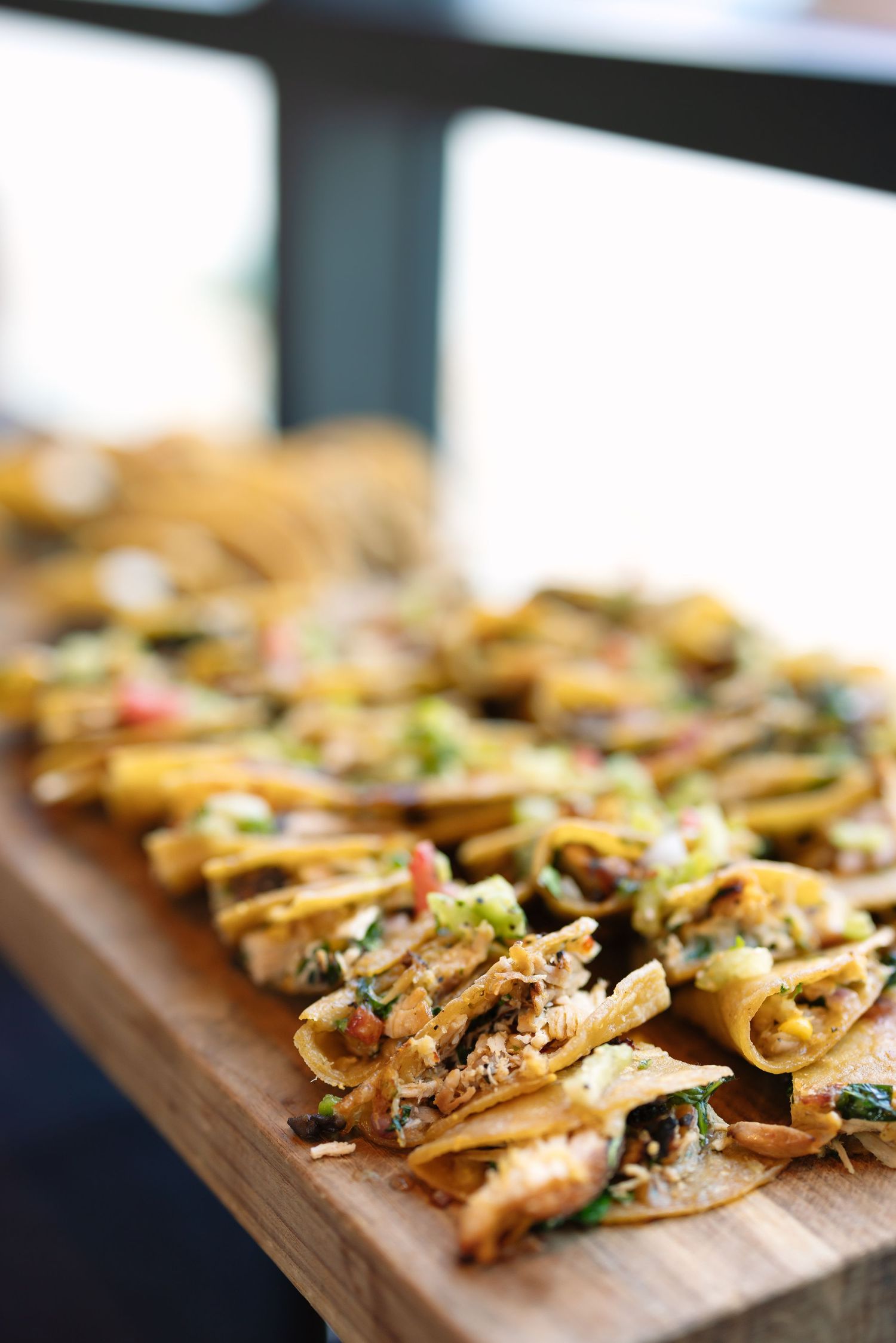 Rows of crispy mini tacos topped with fresh vegetables and herbs served on a wooden board.