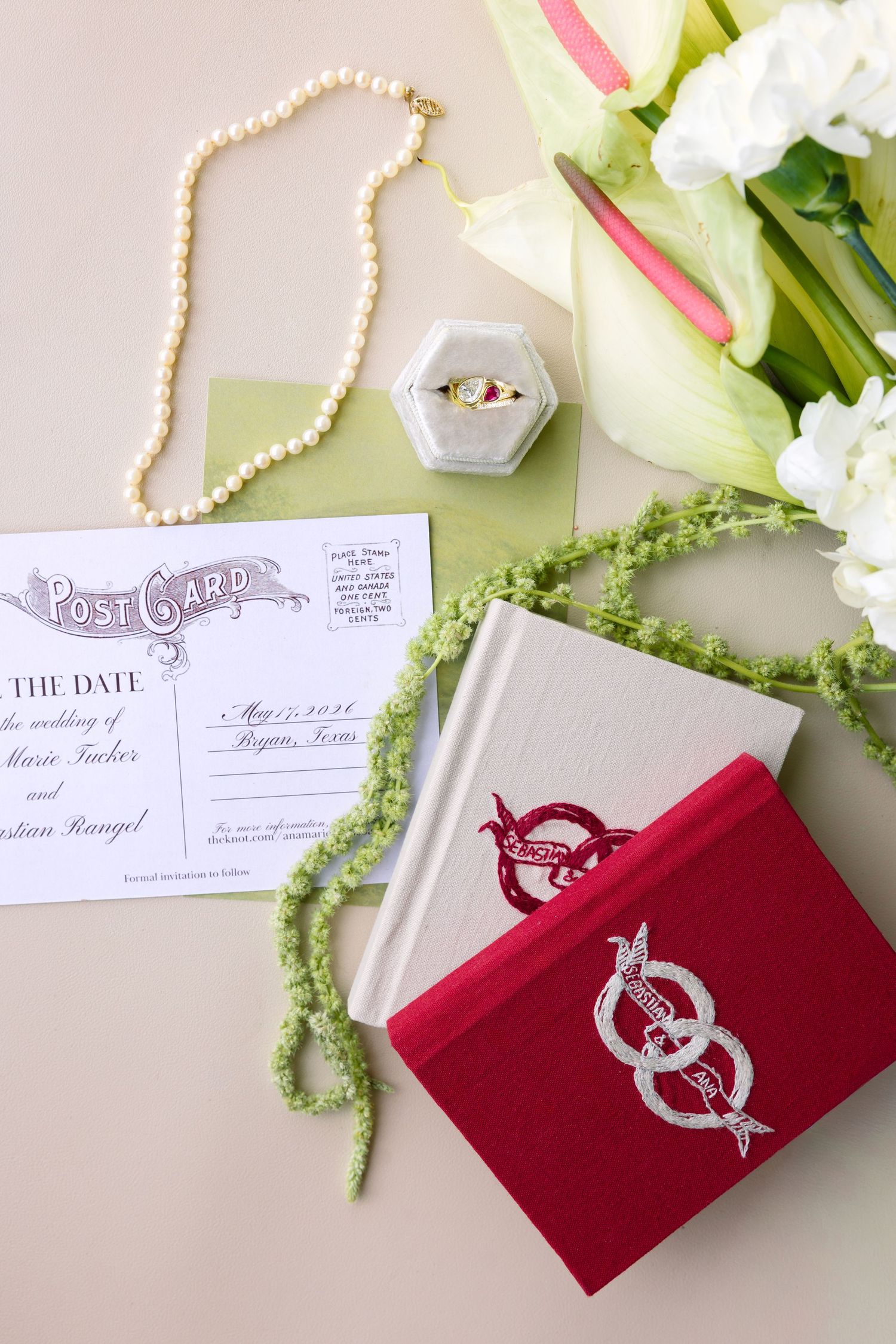 Wedding flat lay with postcard save the date, pearl necklace, ring box, red velvet book, and white floral bouquet.