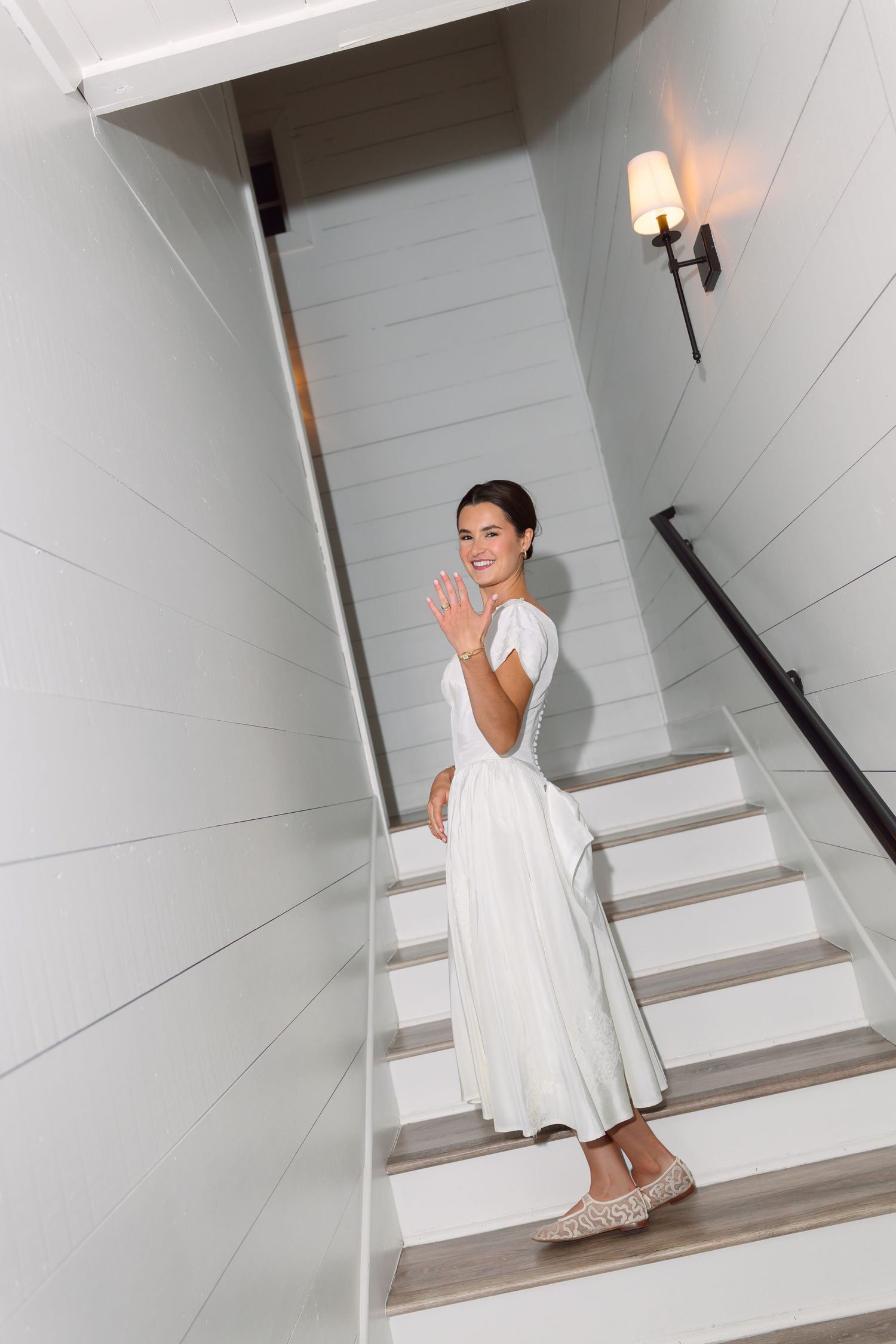 Smiling bride in white tea-length dress waving while descending a staircase in a bright white interior.