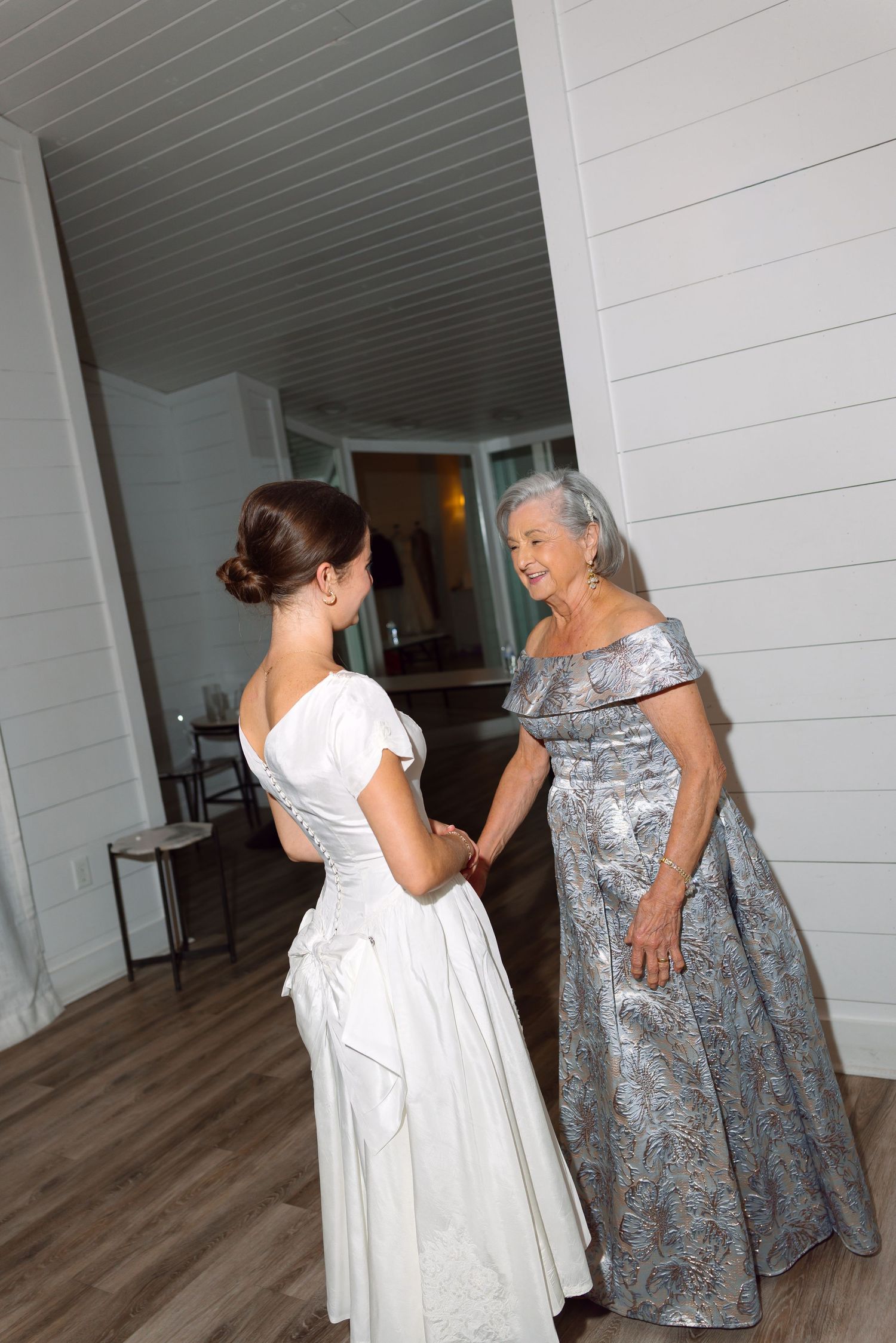 Bride in white gown holds hands with elderly woman in silver dress, sharing a tender moment indoors.