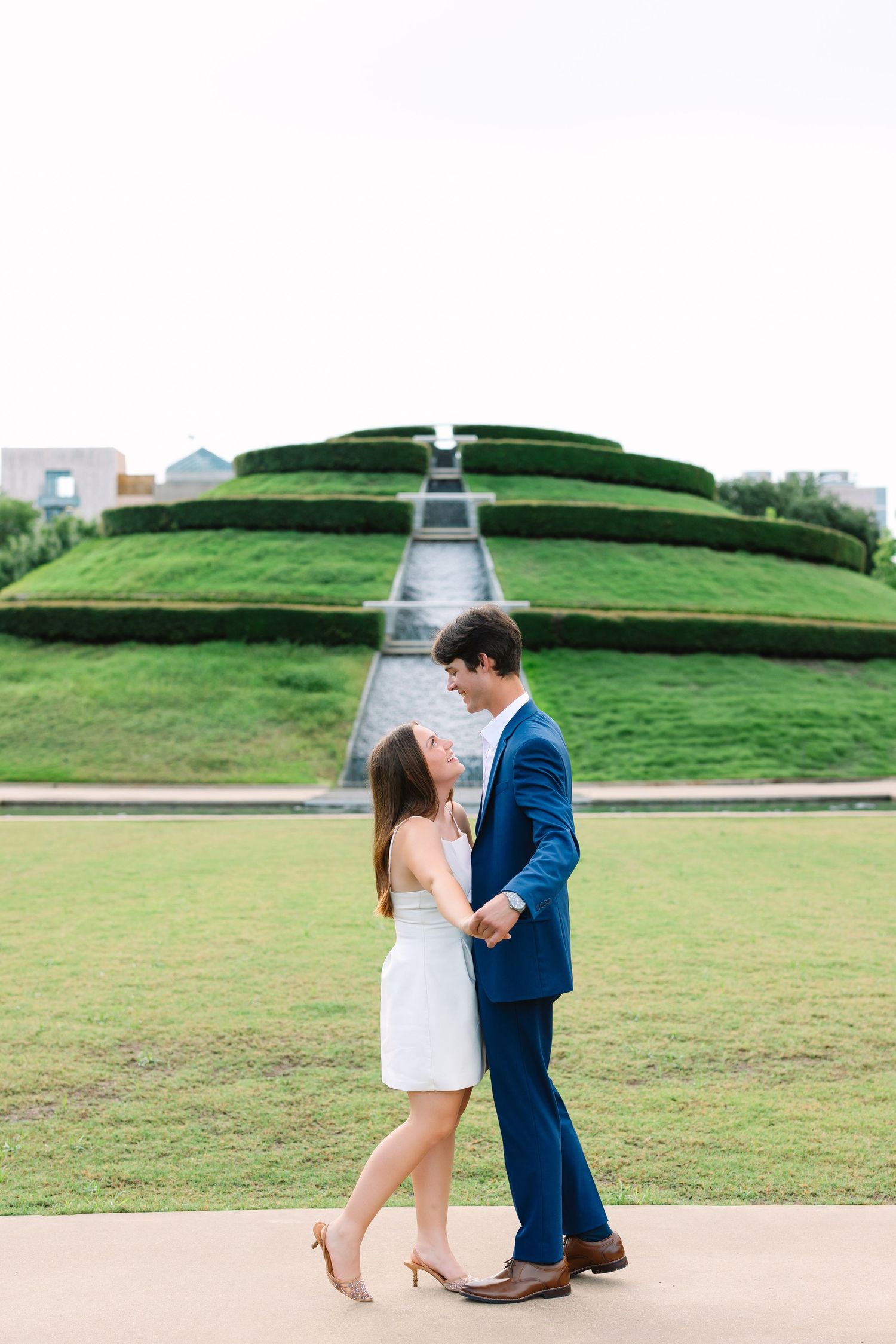 A couple in formal attire poses together on a path with green hills in the background.