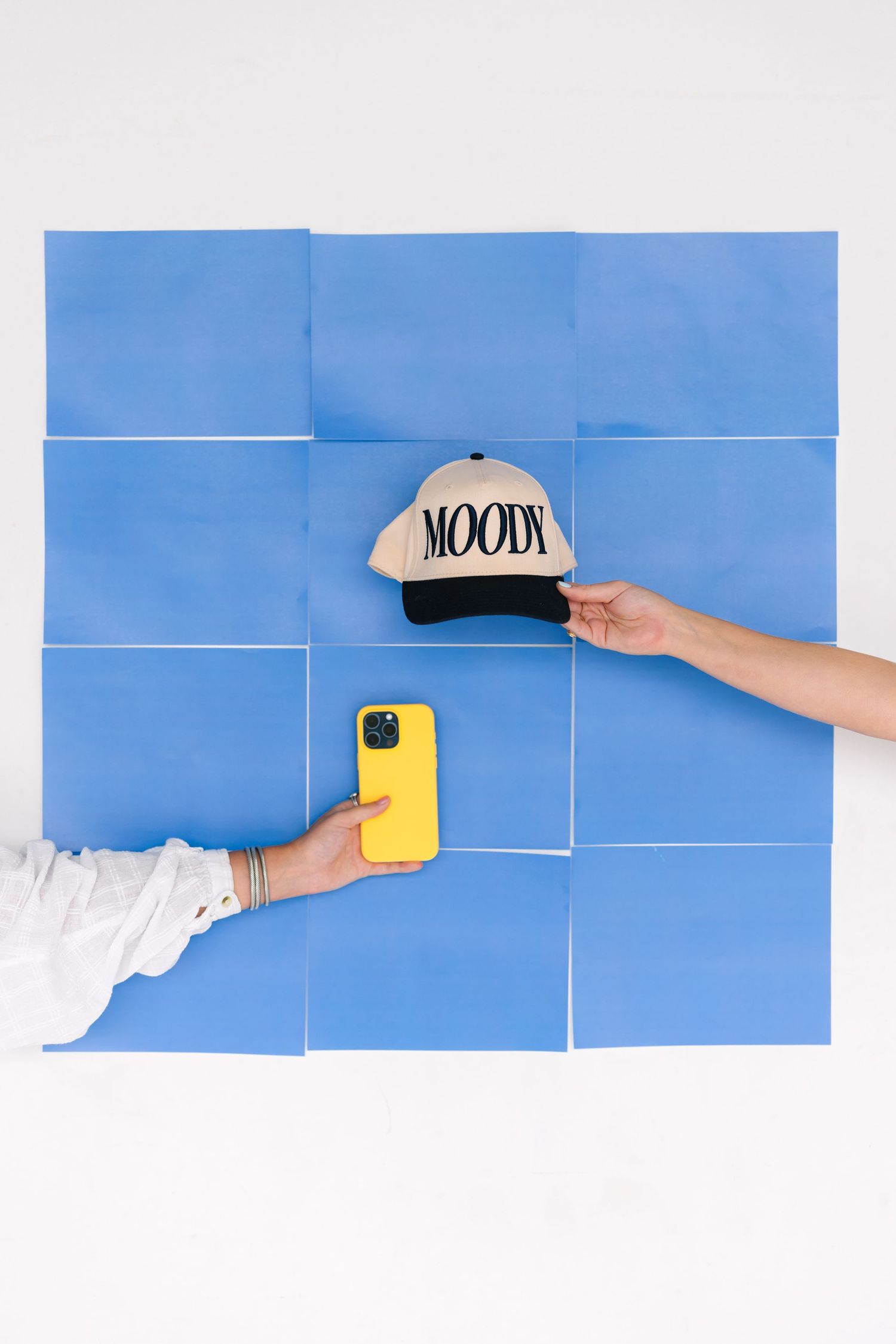 Hands holding a baseball cap and yellow phone against blue geometric wall pattern.