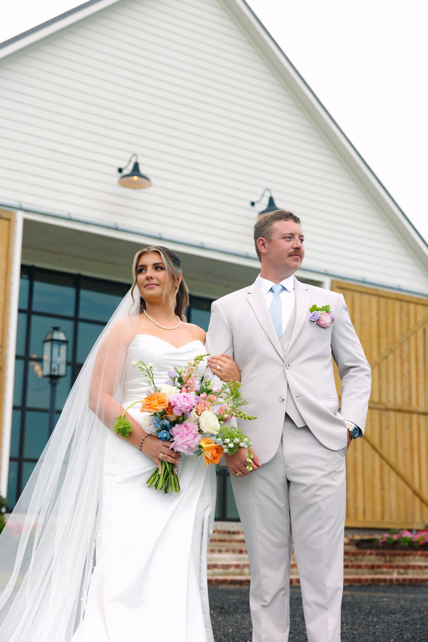 Bride and groom pose together outside a white barn venue, she holds a colorful bouquet wearing a white gown and veil.