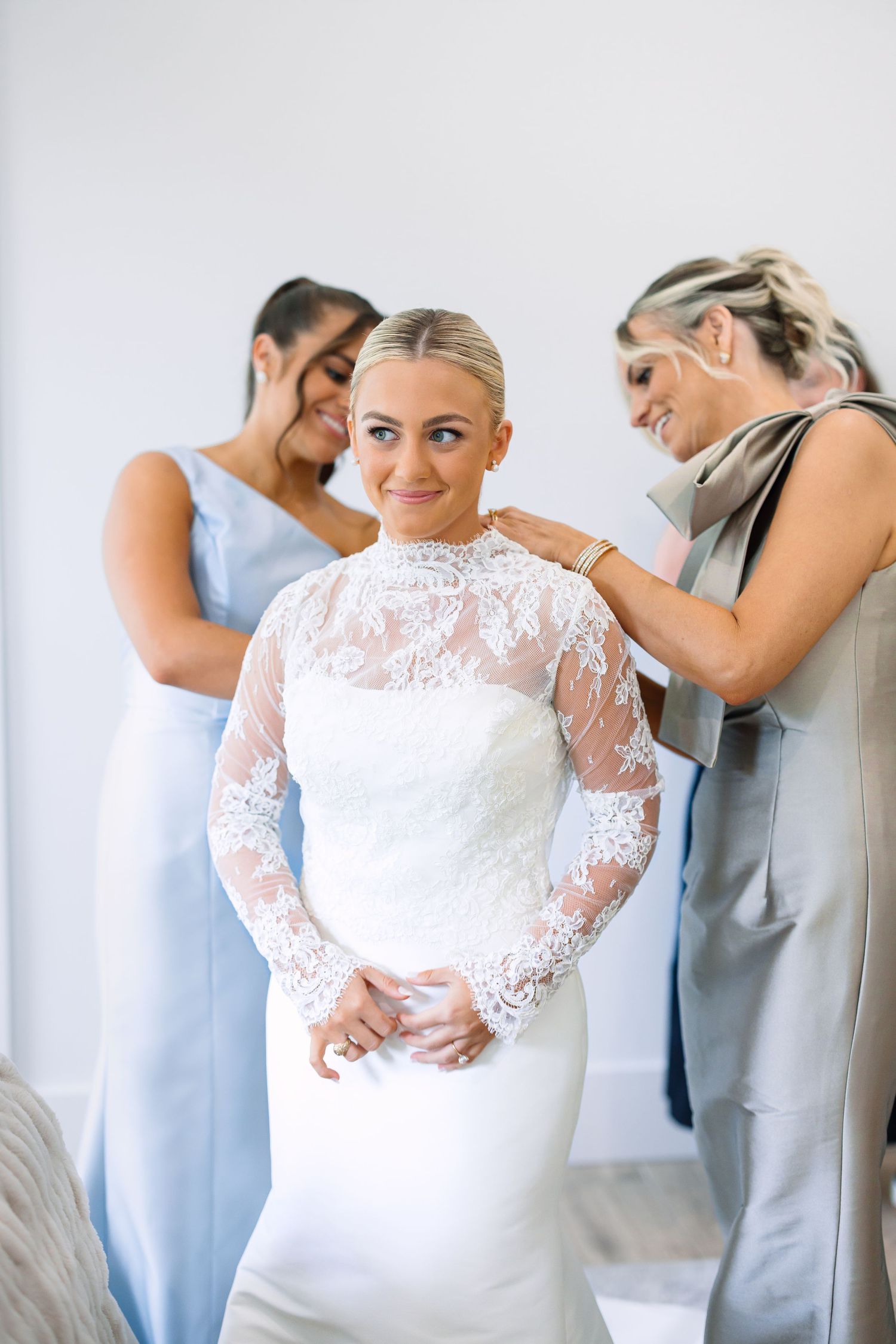 Bride in lace long-sleeve wedding gown smiles as two bridesmaids in blue-gray dresses help her prepare.