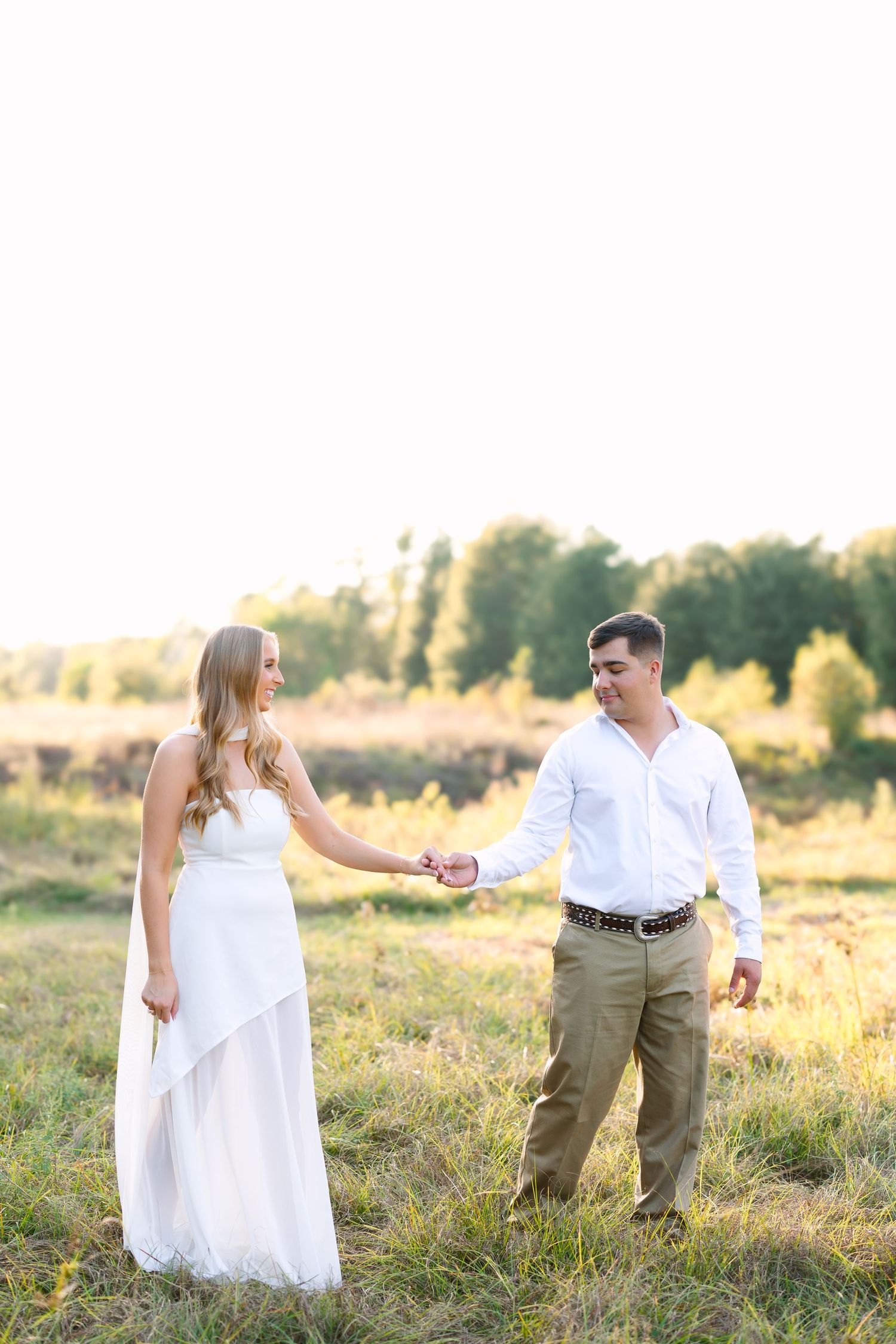 Engaged couple holding hands in a golden sunlit field, woman in white dress and man in white shirt and khaki pants.