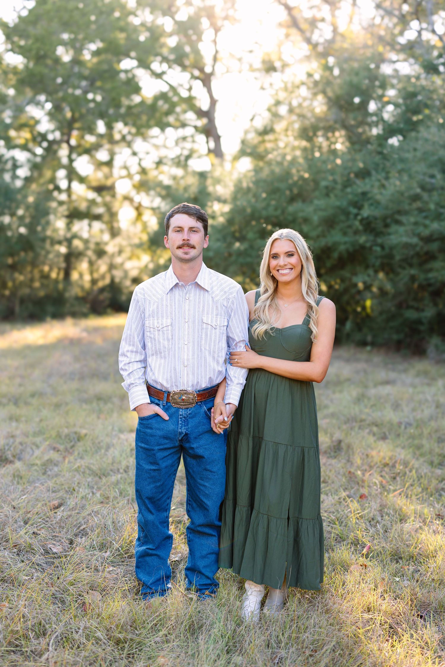 Smiling couple holding hands in golden hour field, woman in green dress and man in striped western shirt.