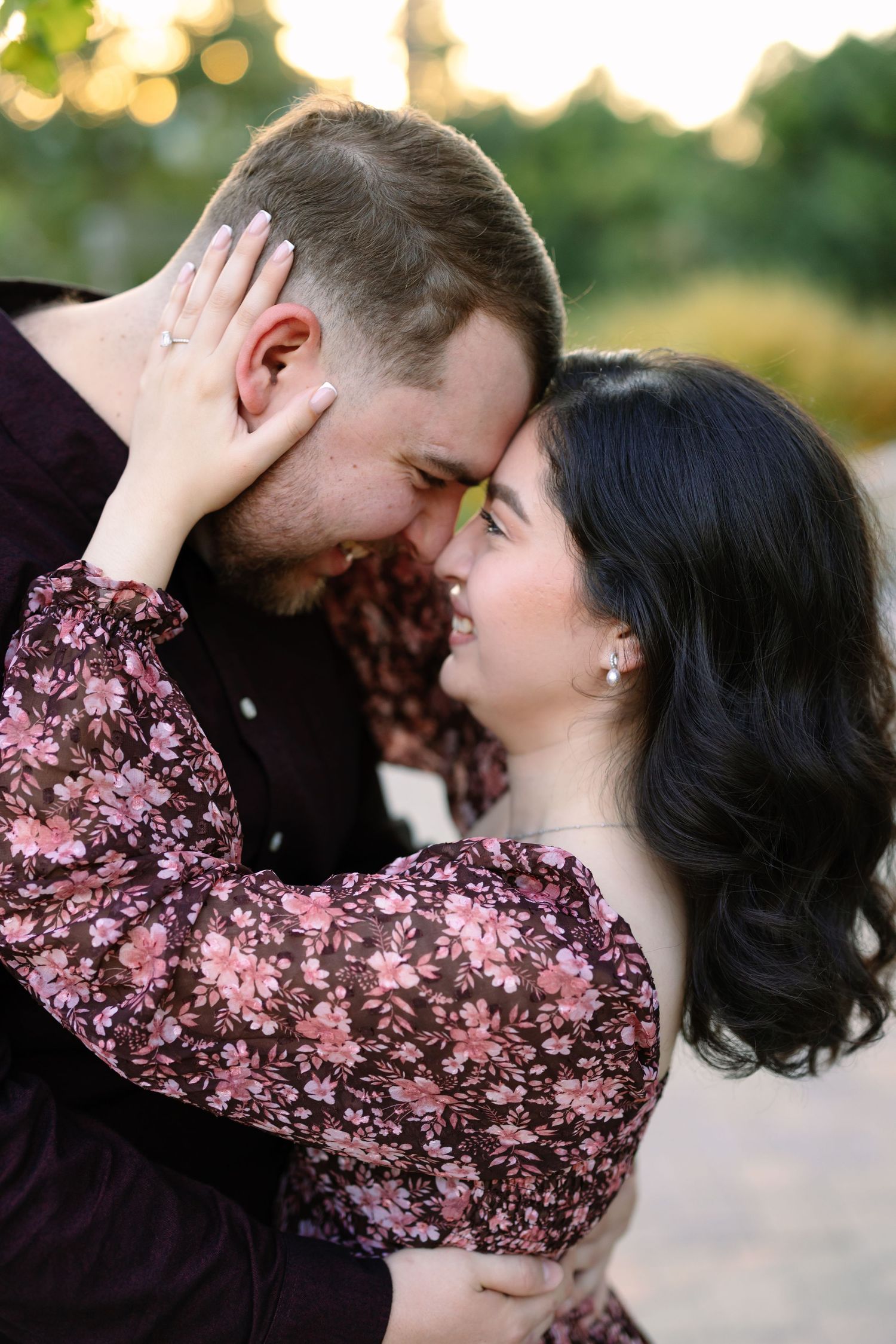 A couple in floral outfits share a romantic forehead touch, smiling tenderly in a softly lit outdoor setting.