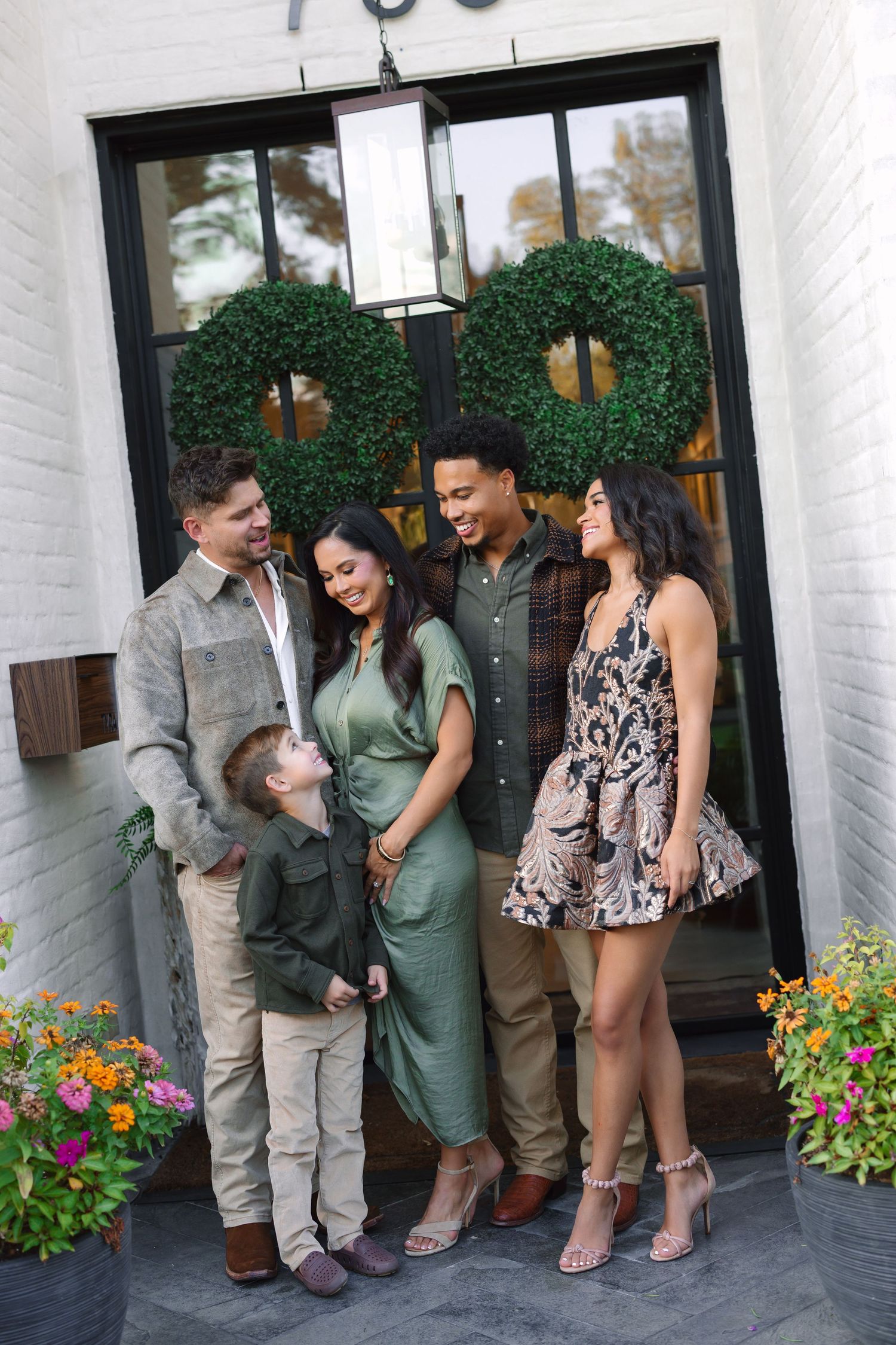 A family of five poses in coordinated green and neutral outfits on a white brick porch with wreath-adorned black doors.