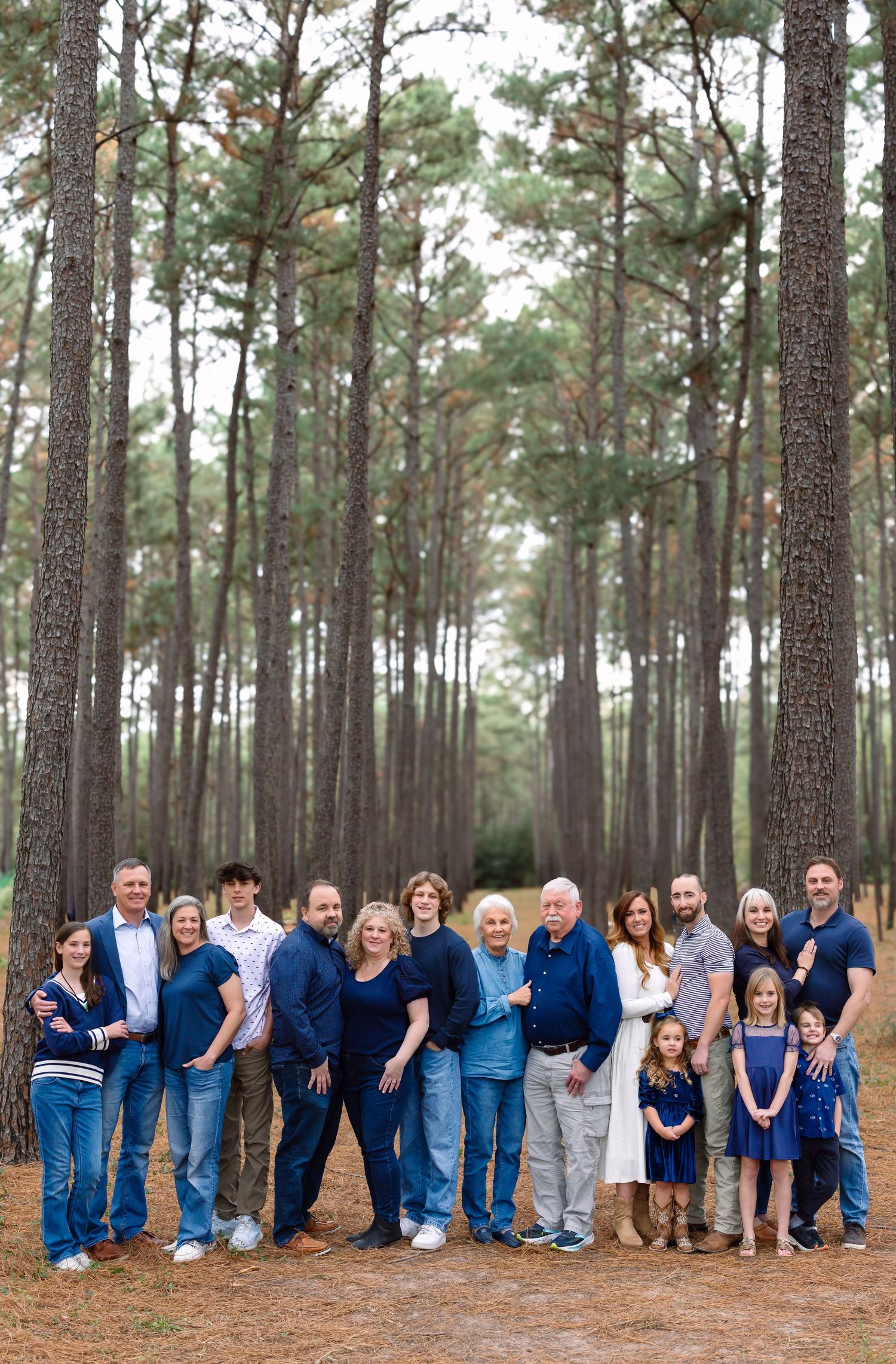 A large multi-generational family dressed in blue tones poses together in a pine tree forest.