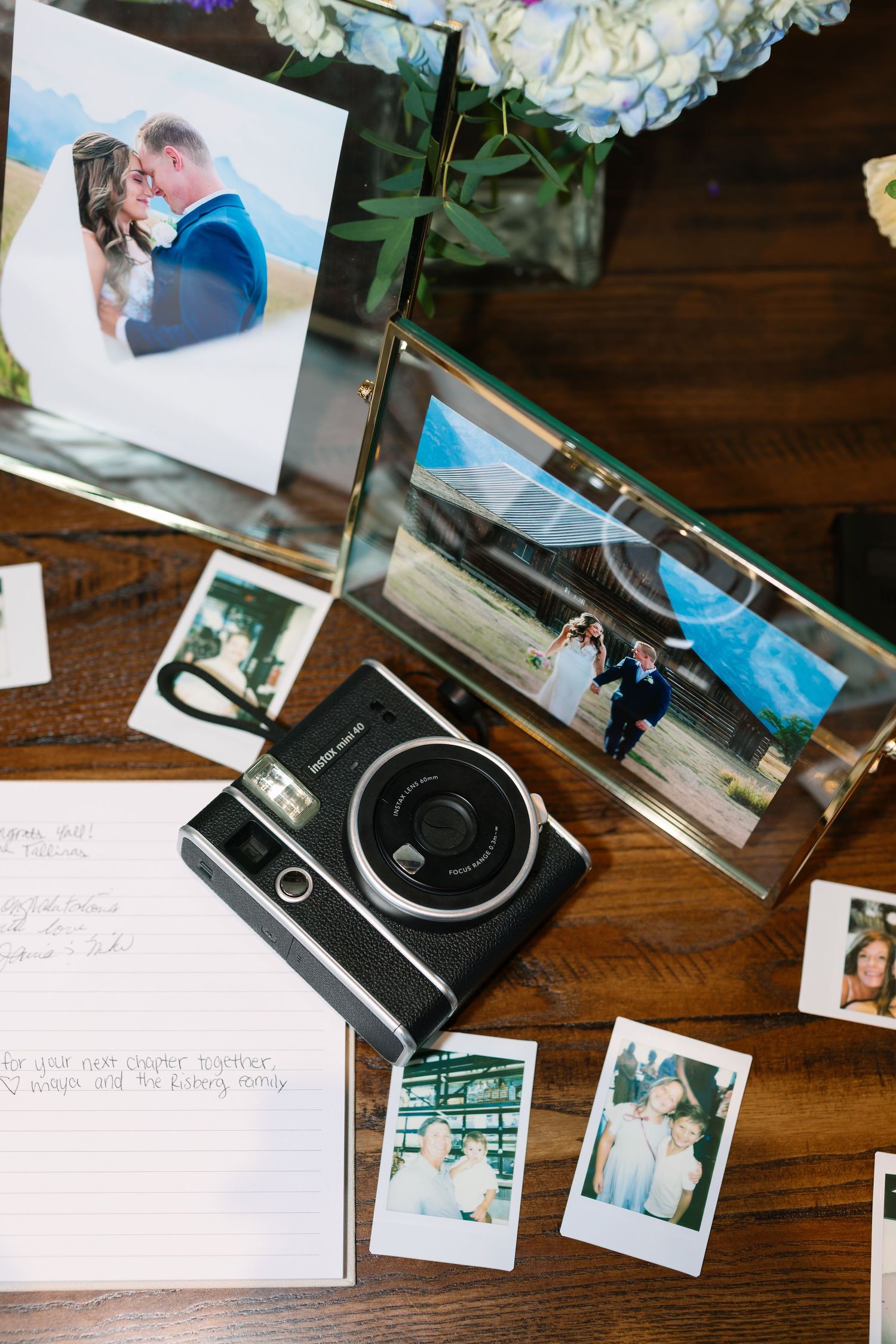 Wedding guest book table with Instax camera, Polaroid photos, handwritten notes, and framed couple portraits on wood surface.