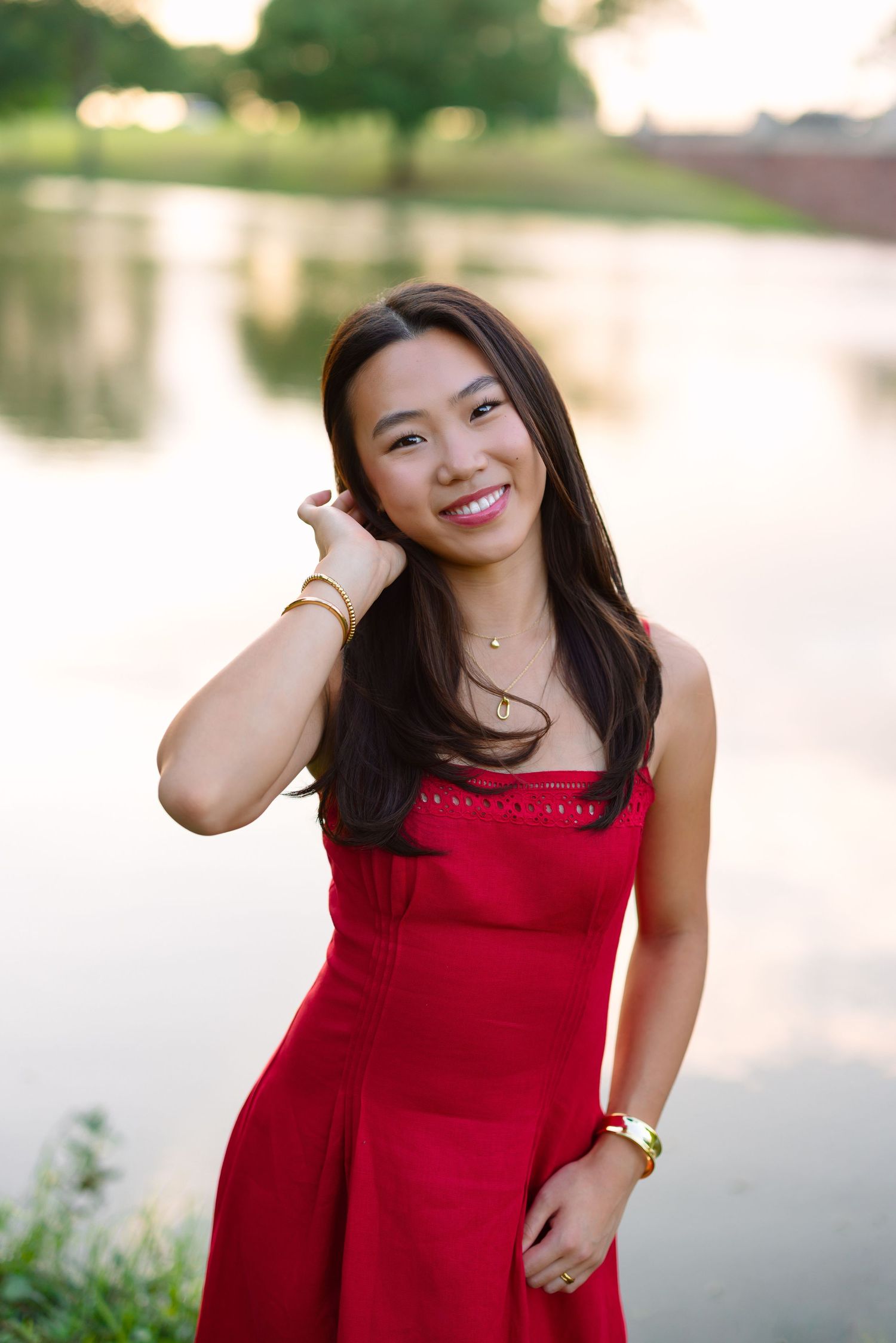 Young Asian woman in red dress smiling outdoors near a serene lake at golden hour.