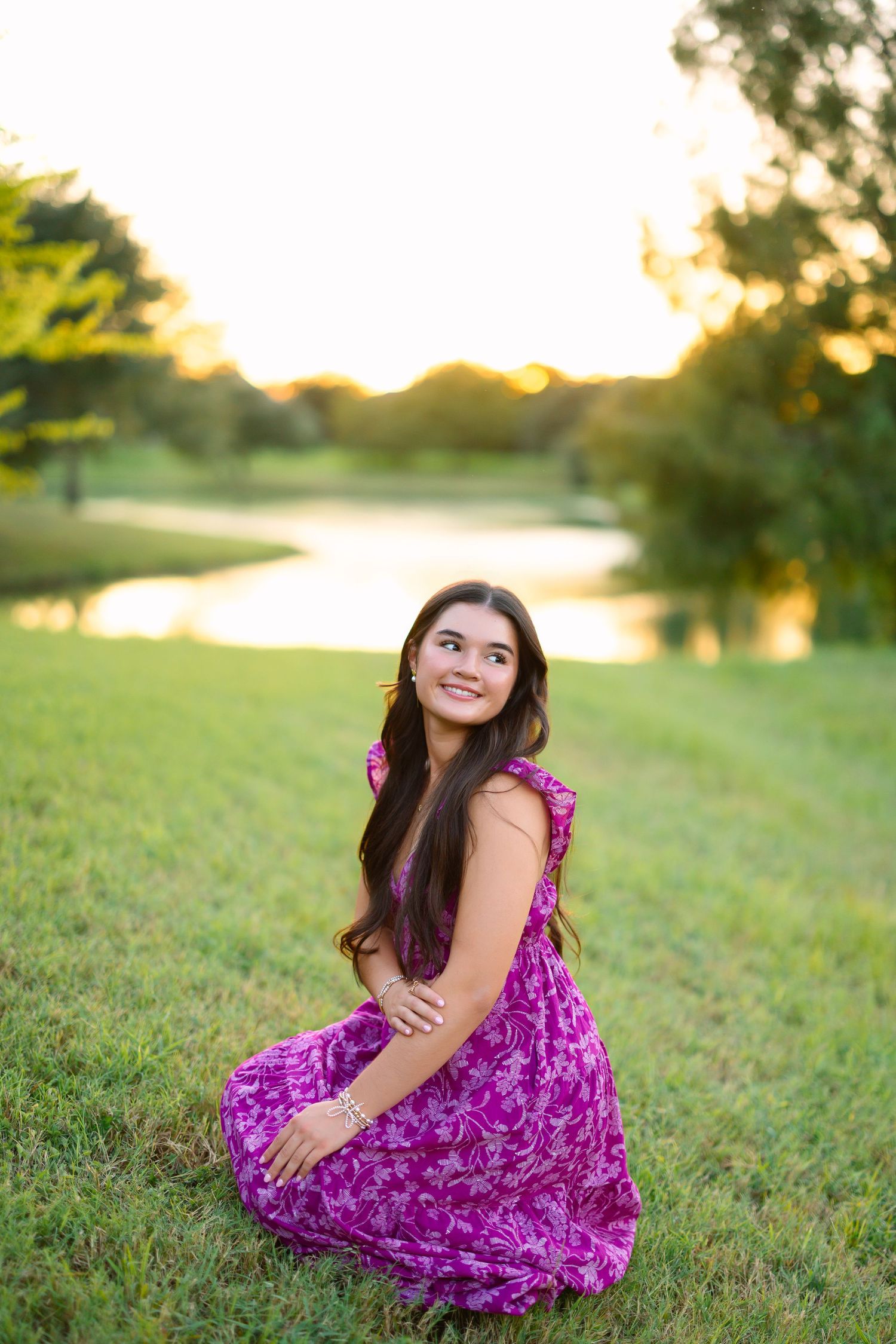 Young woman in purple floral dress sitting gracefully on green grass near a pond during golden hour sunset.