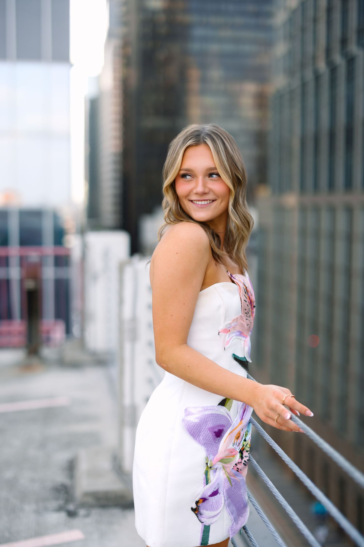 Smiling young woman in a white floral strapless dress poses on an urban rooftop balcony with city buildings in background.