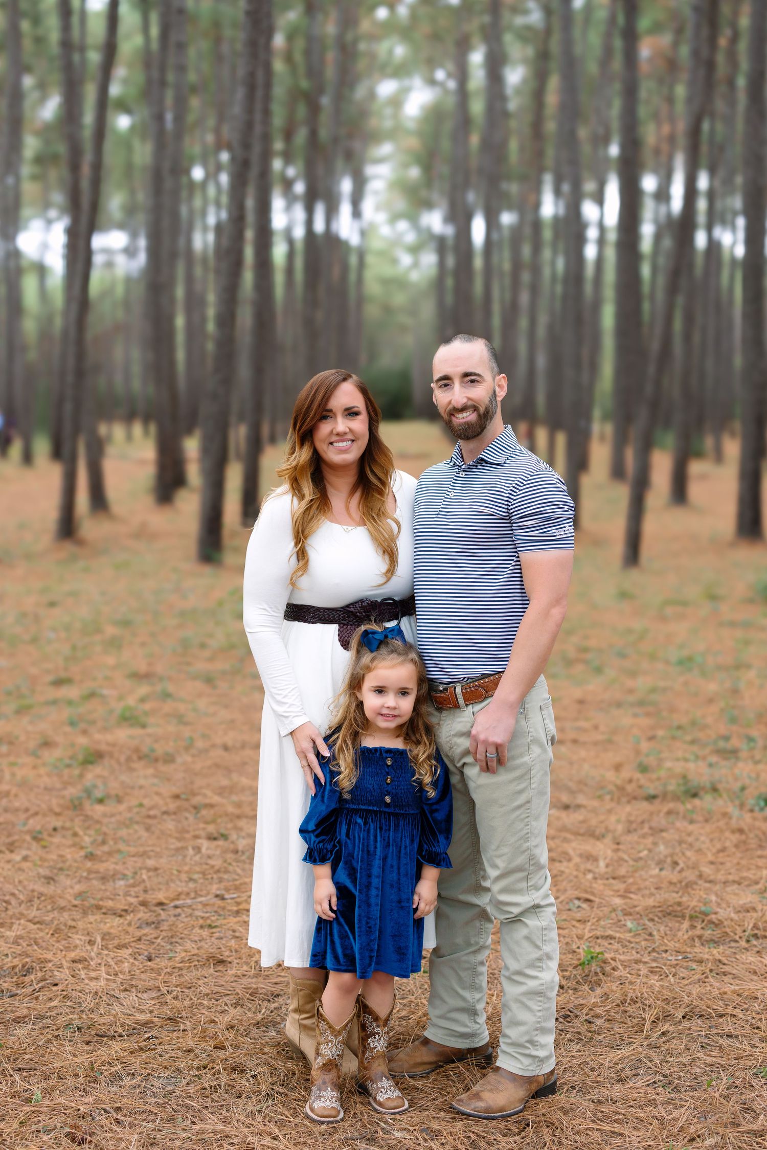 Family of three posing in pine forest, woman in white dress, man in blue shirt, young girl in blue dress.