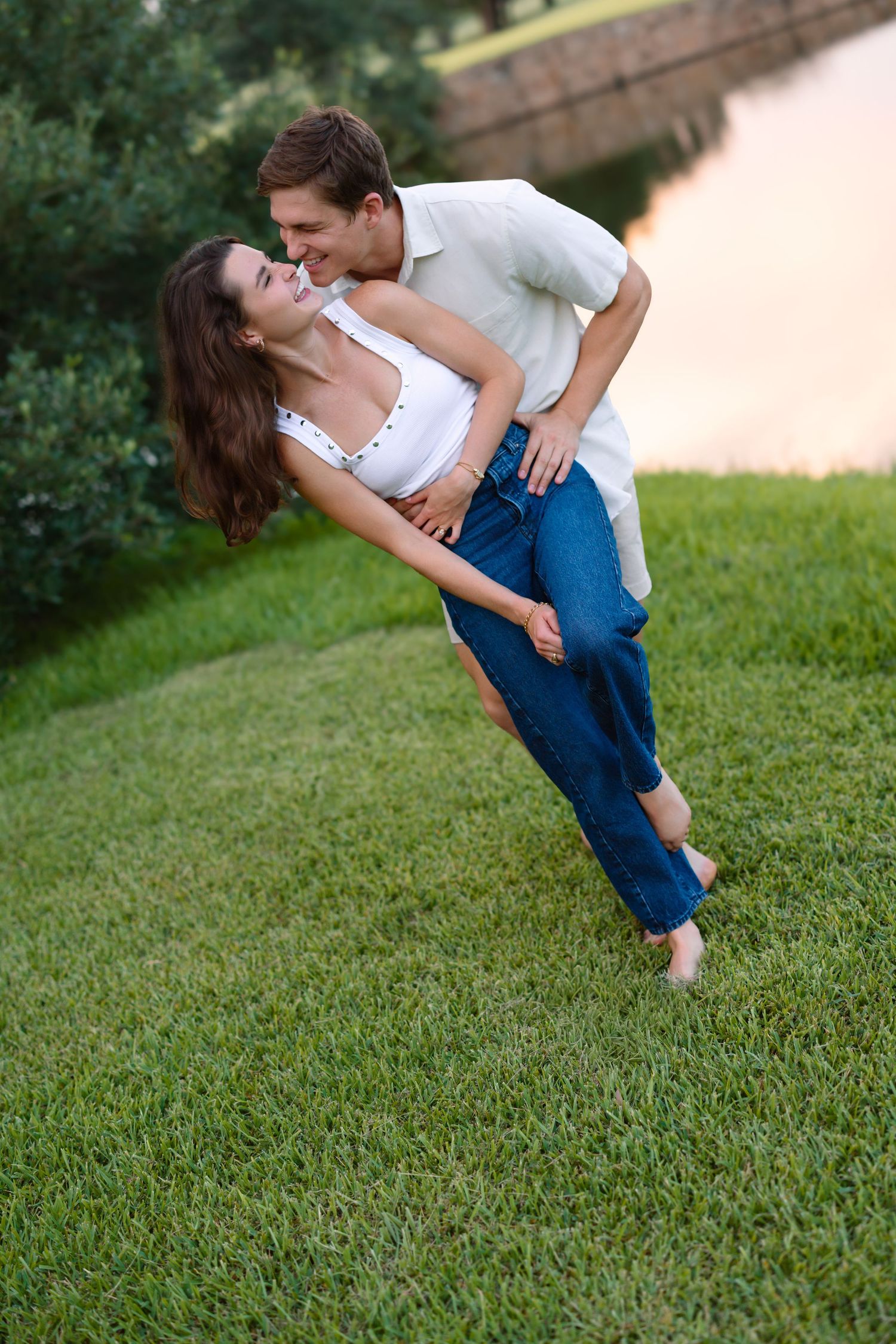 A happy couple playfully embraces on a green lawn near a pond, with the man lifting the woman in jeans and white top.
