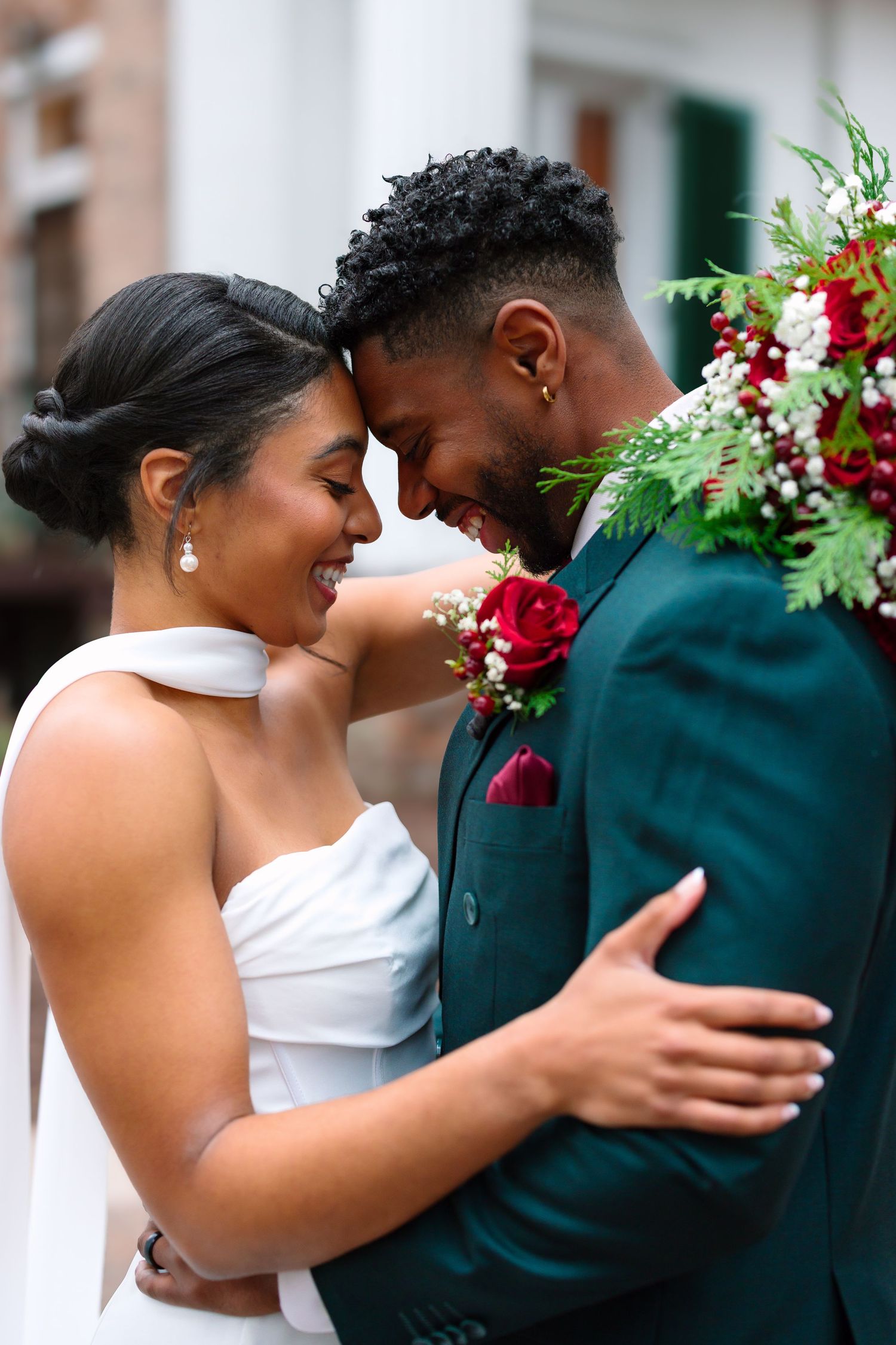 Smiling bride and groom in green suit with red rose boutonniere touching foreheads in a romantic wedding portrait.