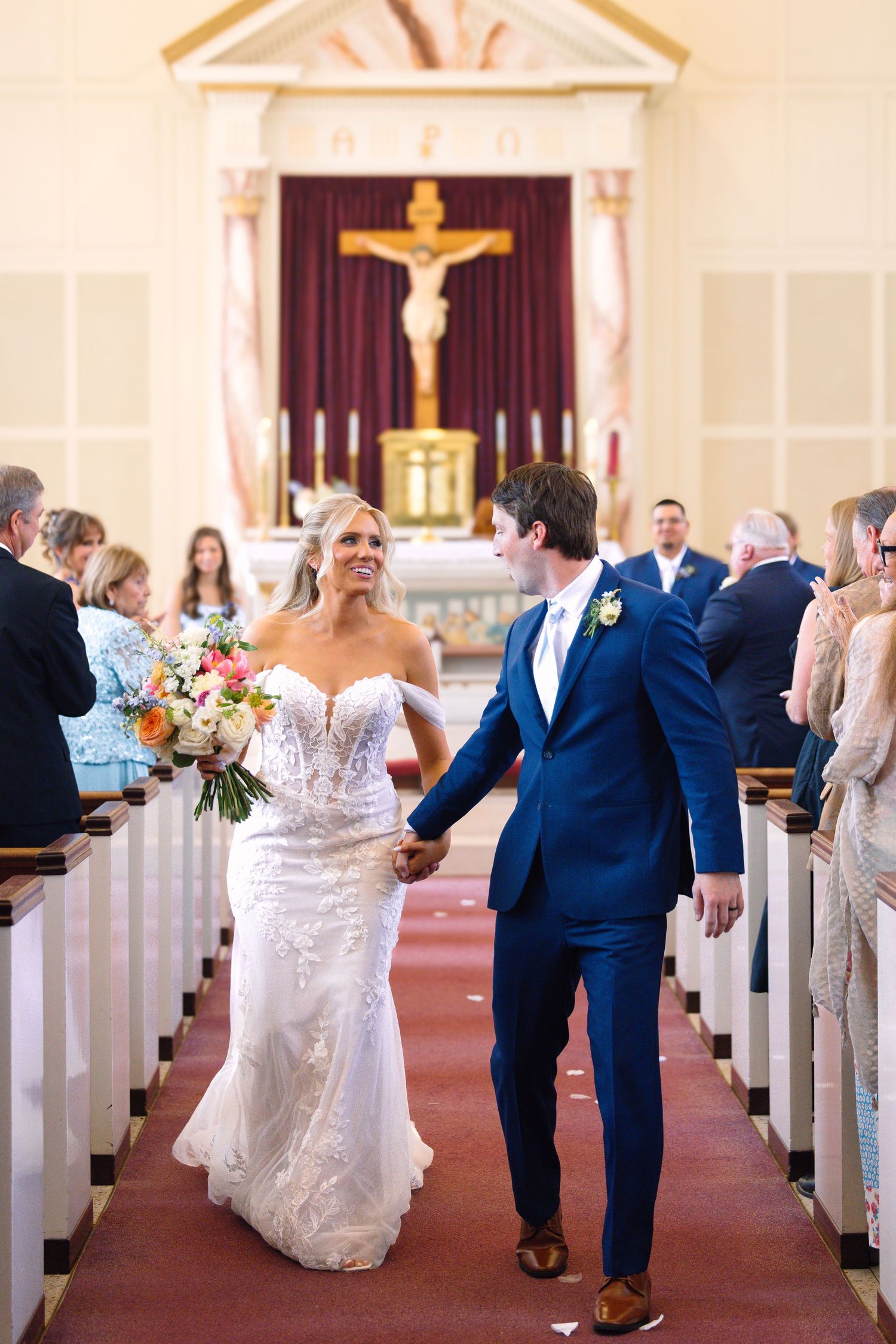 Bride and groom walk down church aisle after wedding ceremony, bride holding colorful bouquet, groom in navy suit.