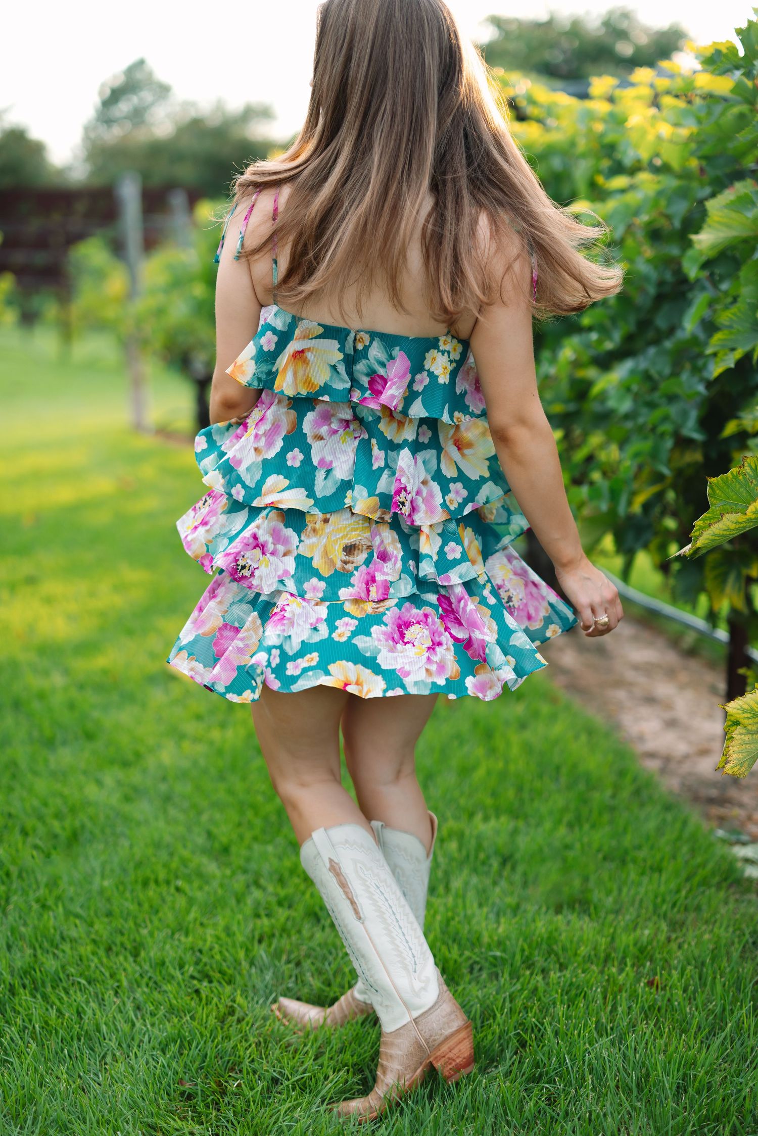 Young child in a floral dress and white boots twirling playfully on green grass.