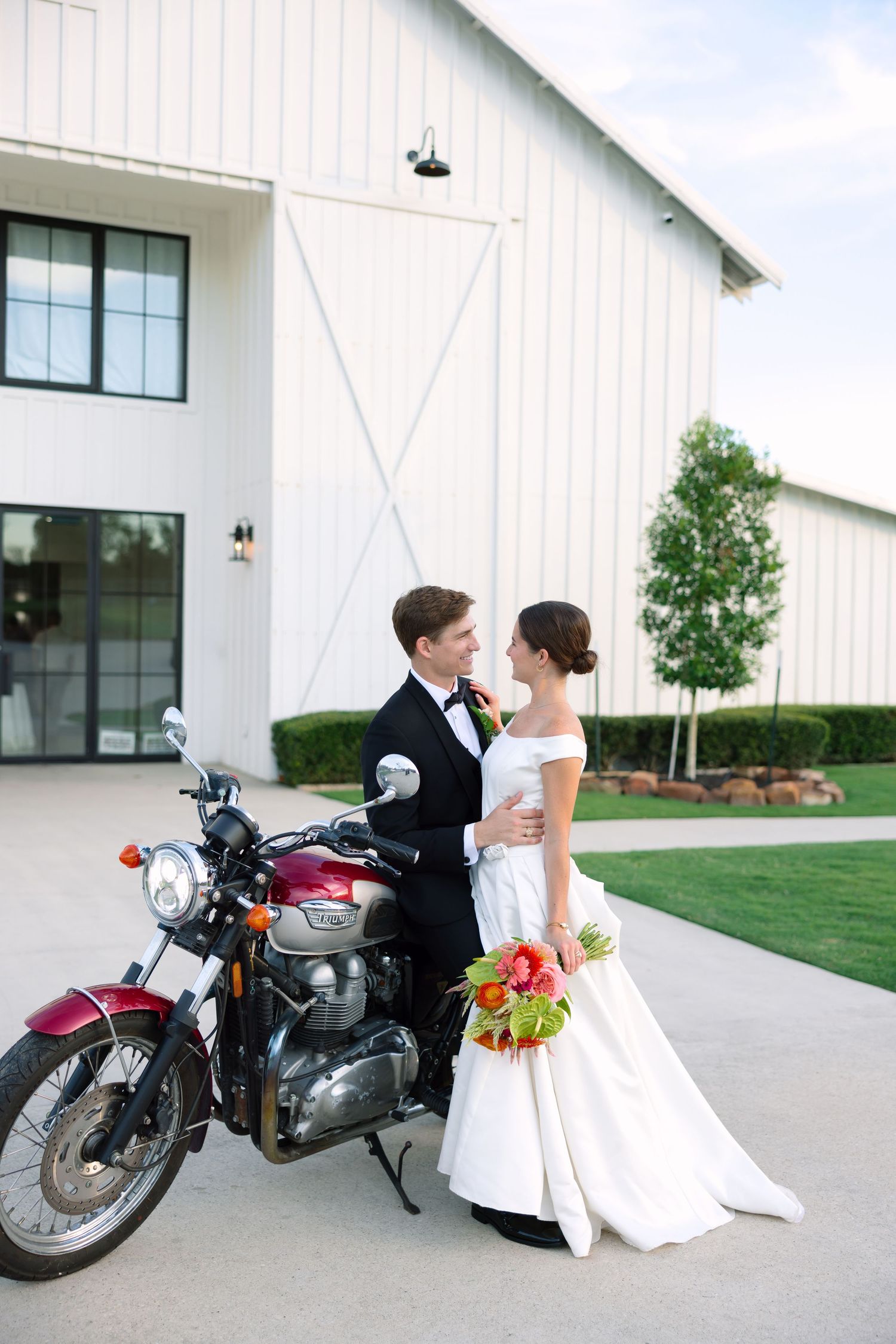 Bride and groom pose together beside a vintage red motorcycle in front of a white barn venue.