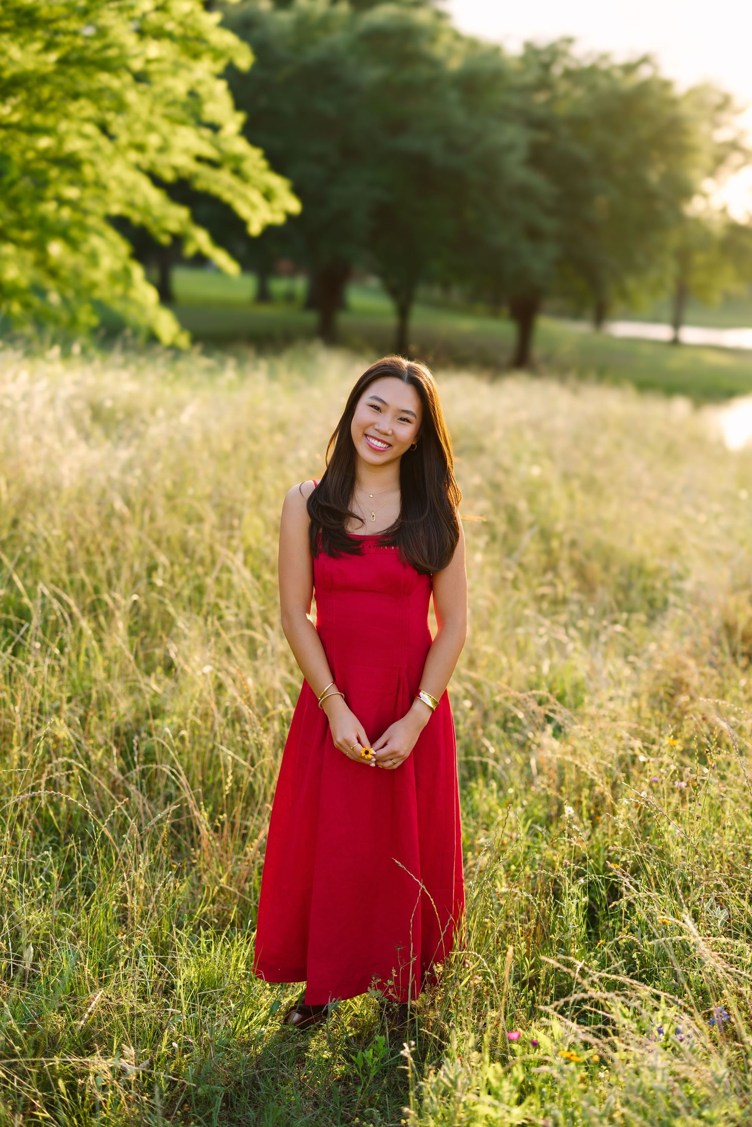 Young Asian woman in red maxi dress smiling in sunlit meadow with green trees in background.