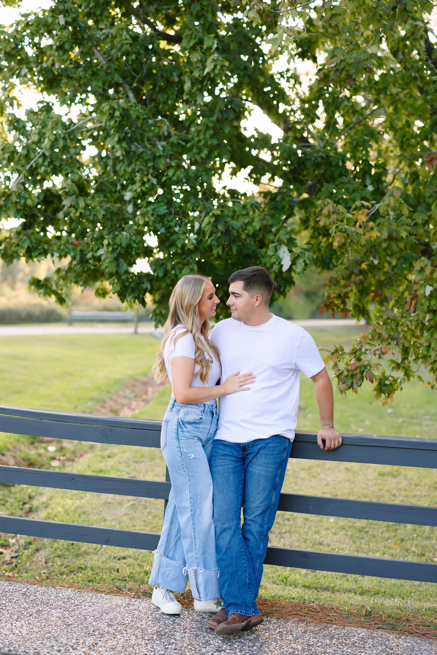 Couple in casual white and denim outfits gazing at each other by a dark fence with lush green trees behind.