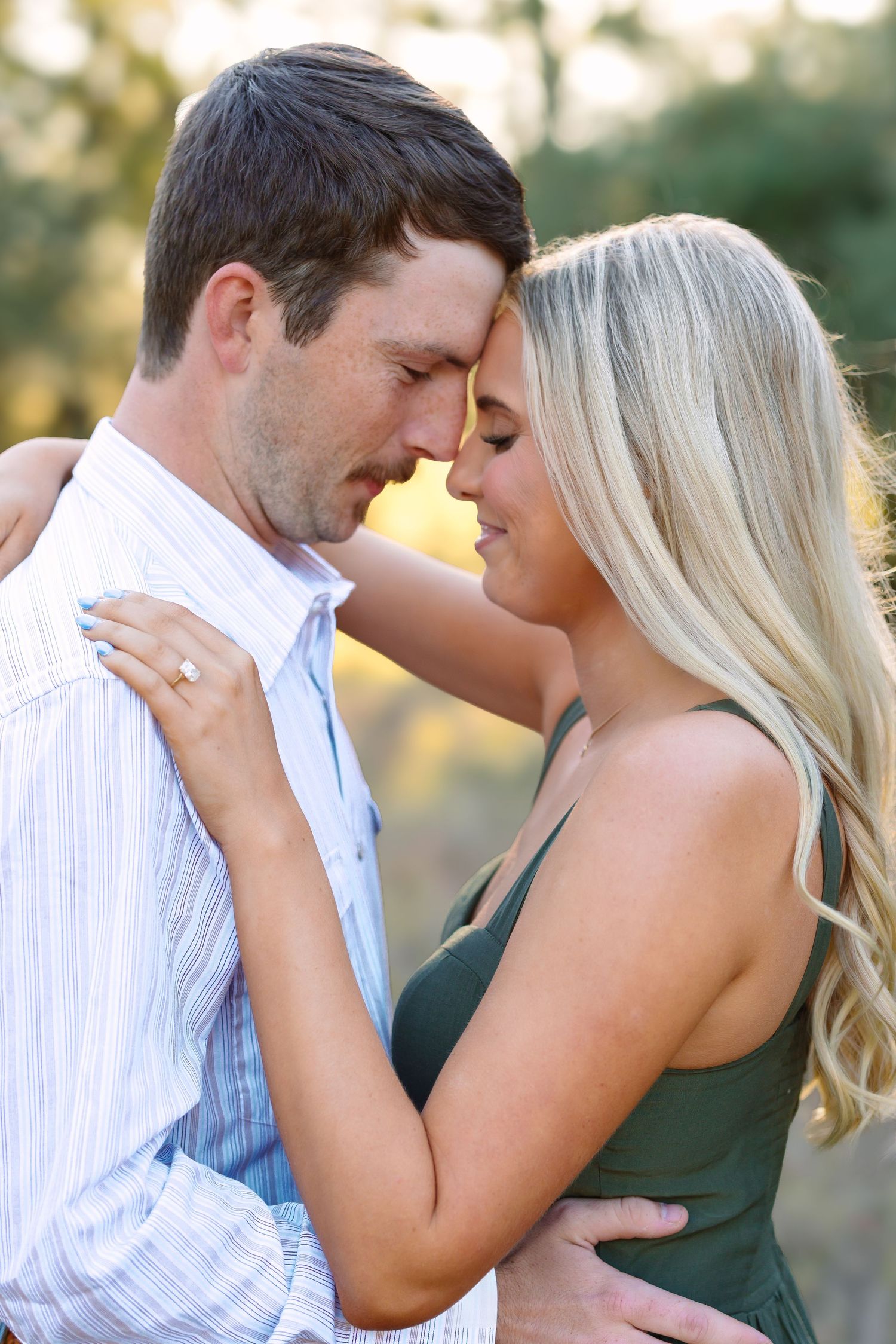 A couple shares a romantic forehead-to-forehead moment outdoors, the woman wearing an engagement ring and green dress.