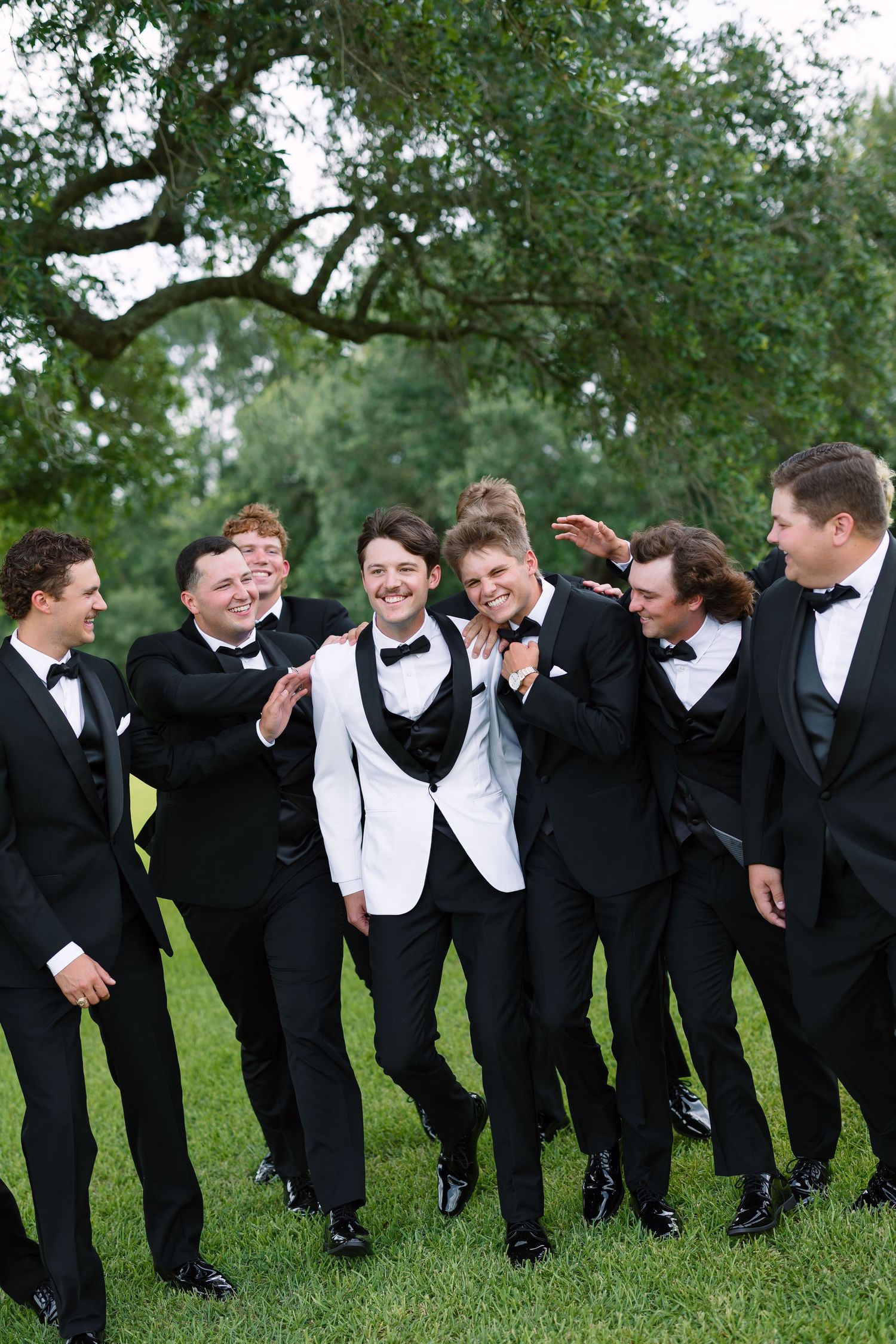 Groom in white tuxedo jacket laughing with groomsmen in black tuxedos outdoors under oak trees.