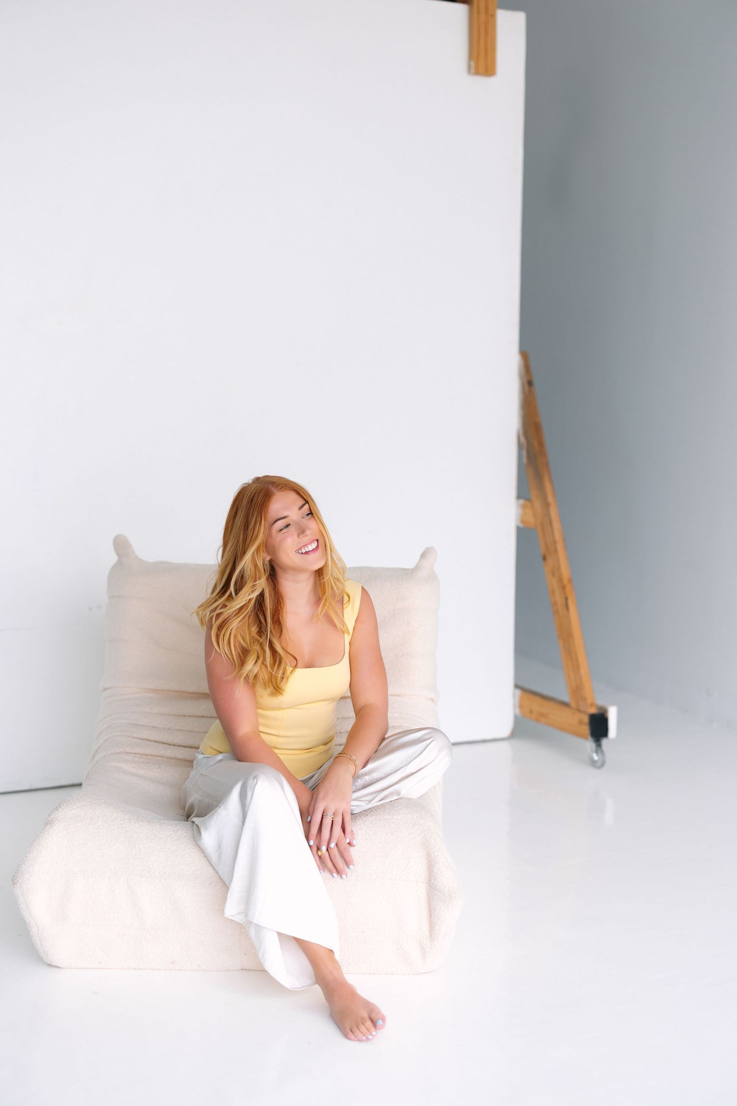 Person in yellow top sitting relaxed on white cushions in minimal studio setting.