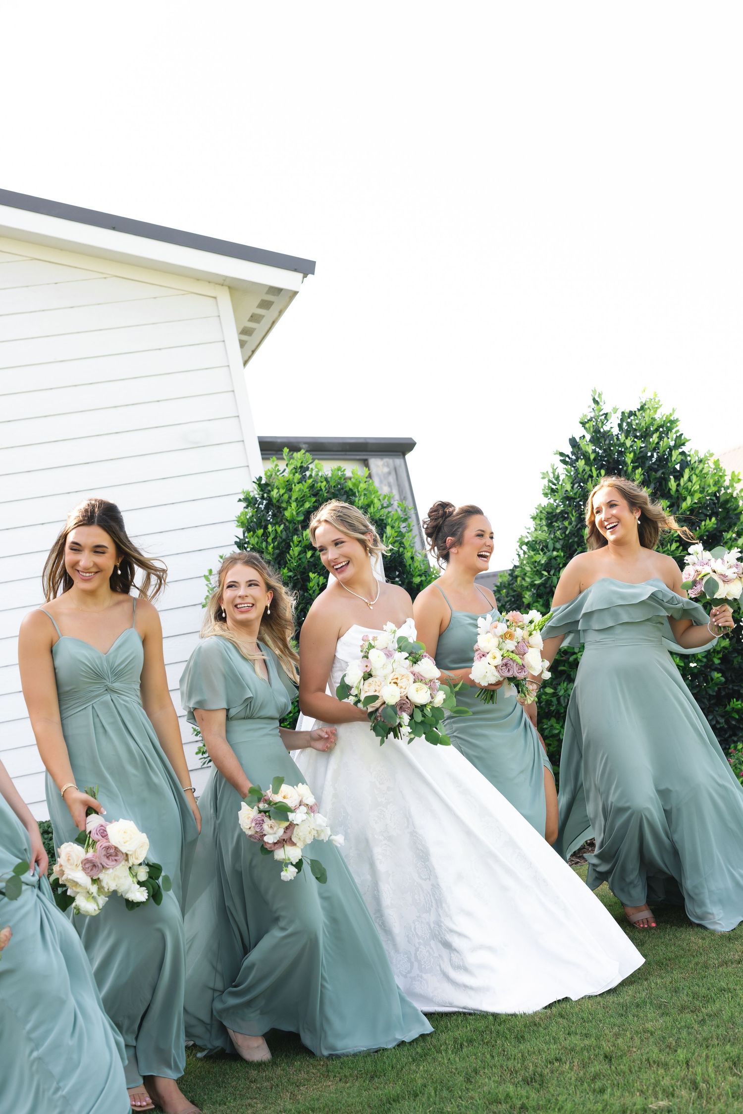 Bridesmaids in sage green dresses pose playfully on grass near white building.