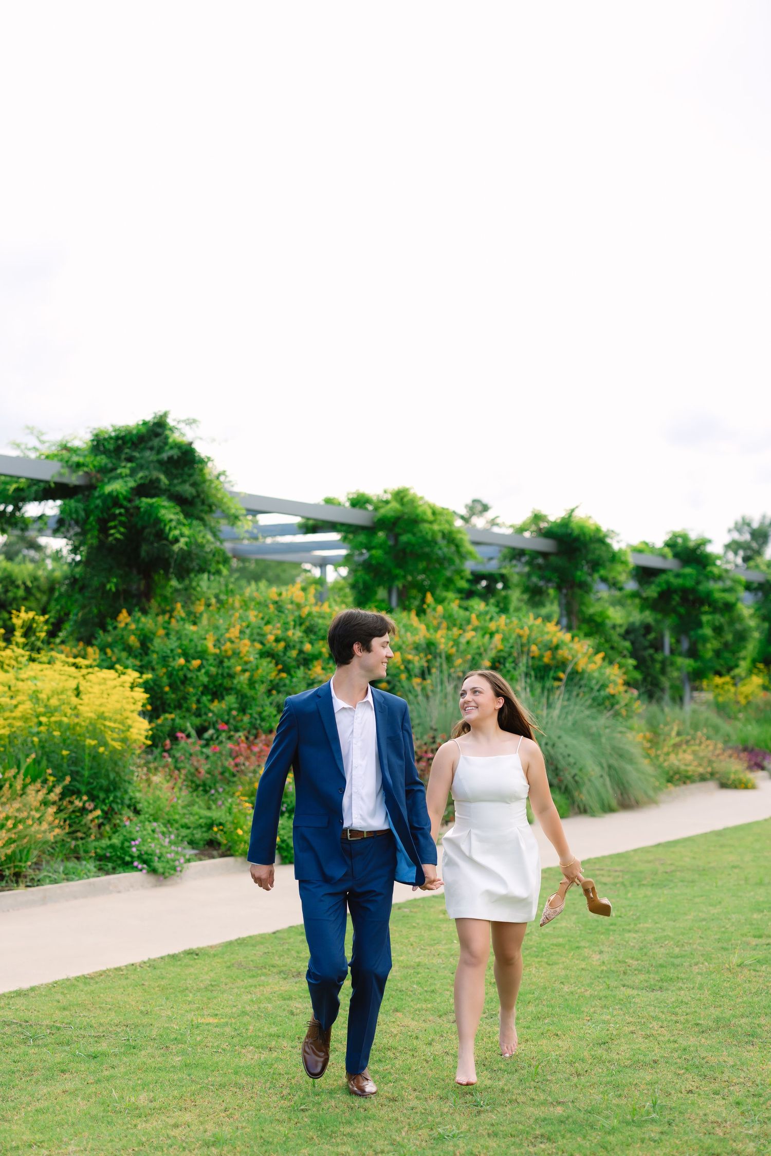 A couple in formal attire walks hand in hand along a garden path surrounded by lush greenery.