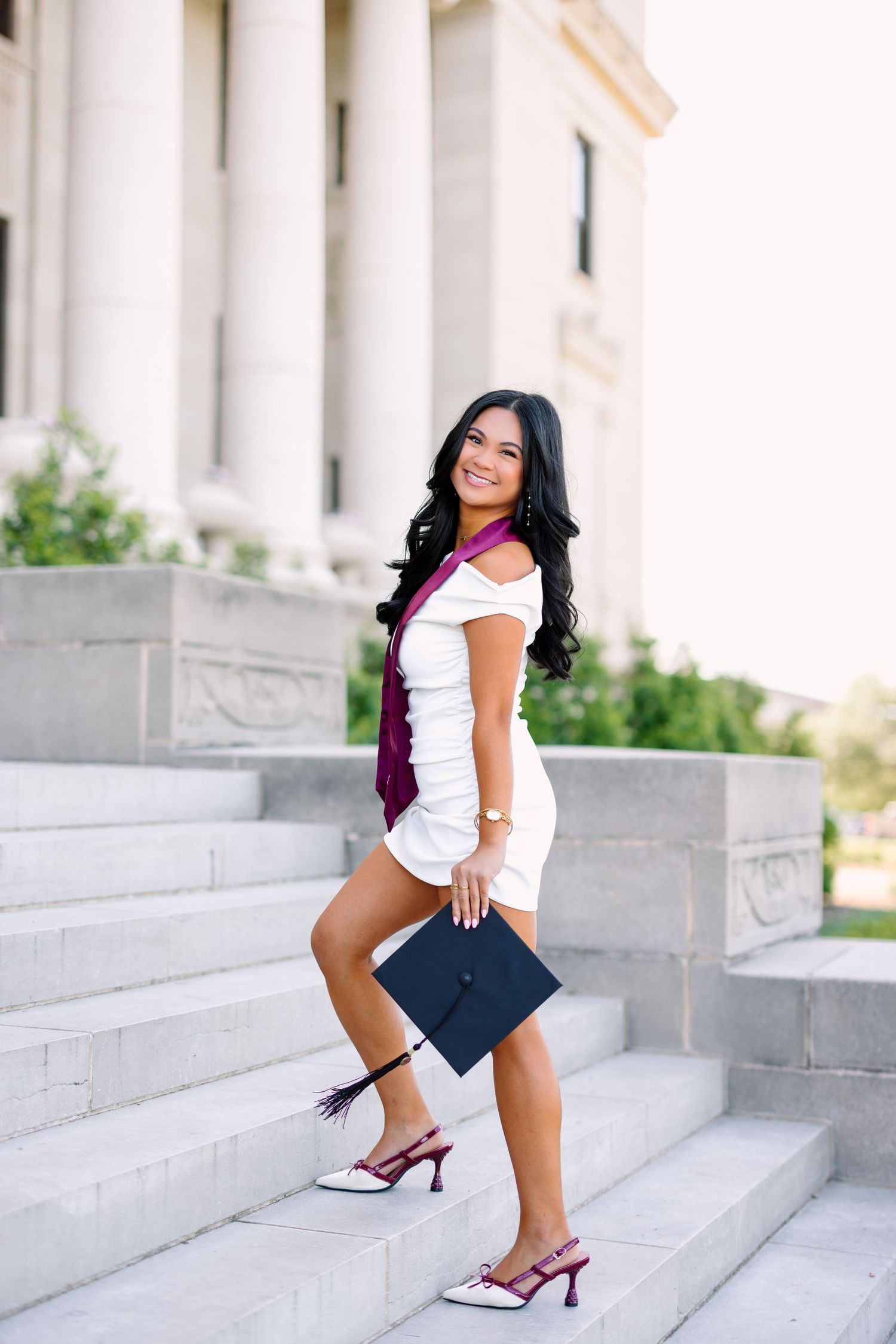 Graduate in cap and gown poses on white marble steps during graduation photoshoot.